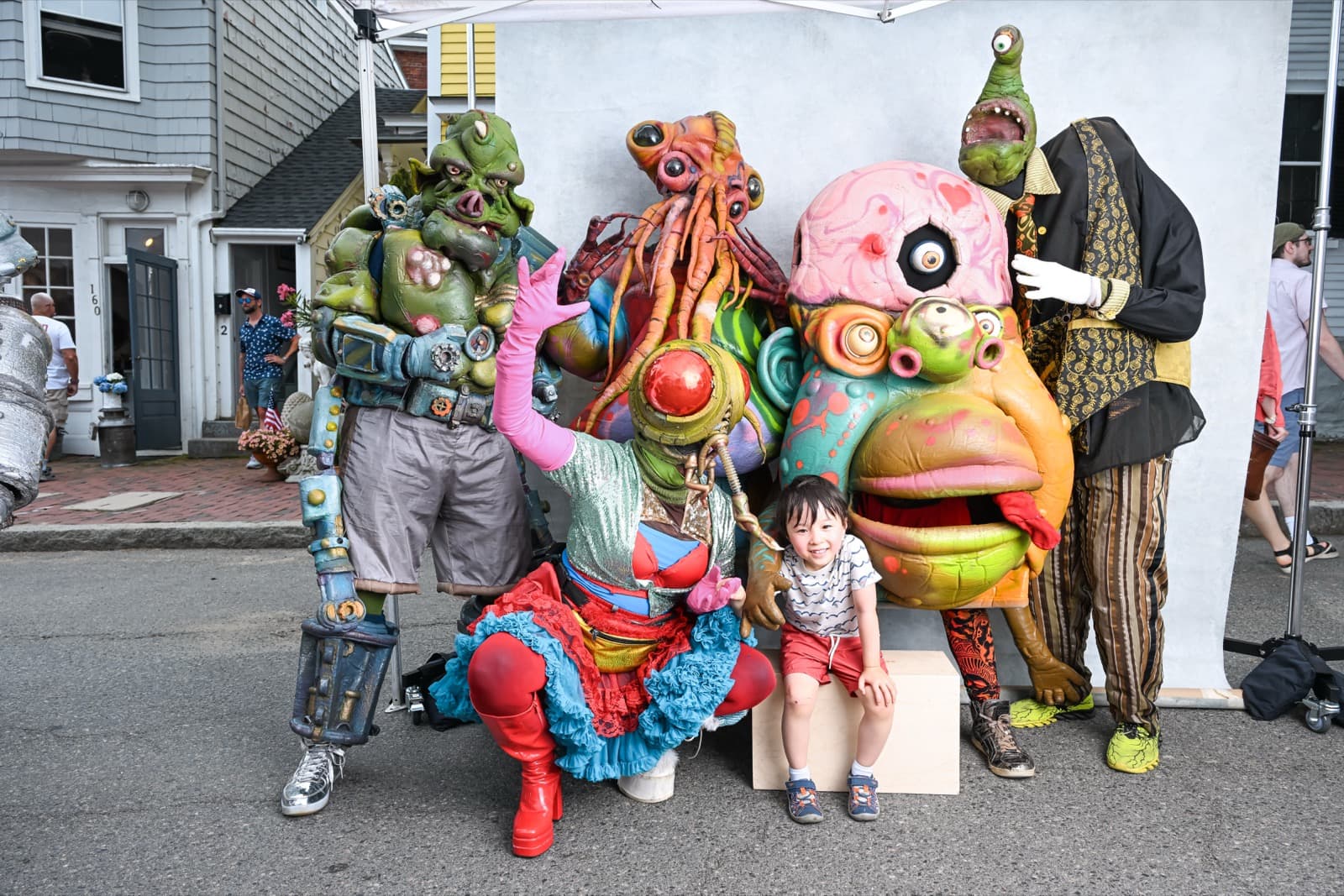 Child sitting on a wooden block, laughing with a group of elaborate monster and creature costume characters.