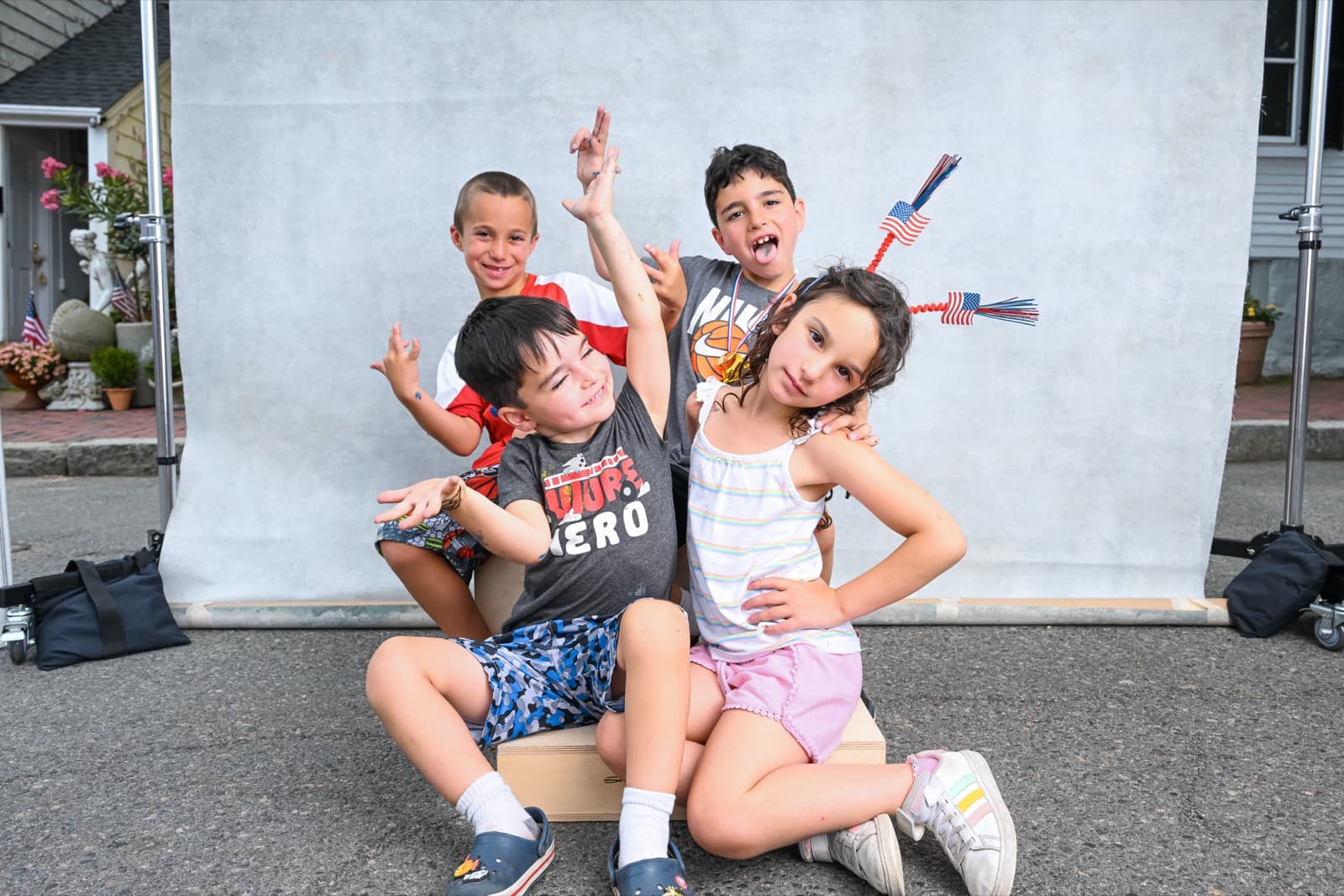 Four kids making playful faces in front of a portable photo backdrop, one boy wearing a basketball medal and patriotic headband.
