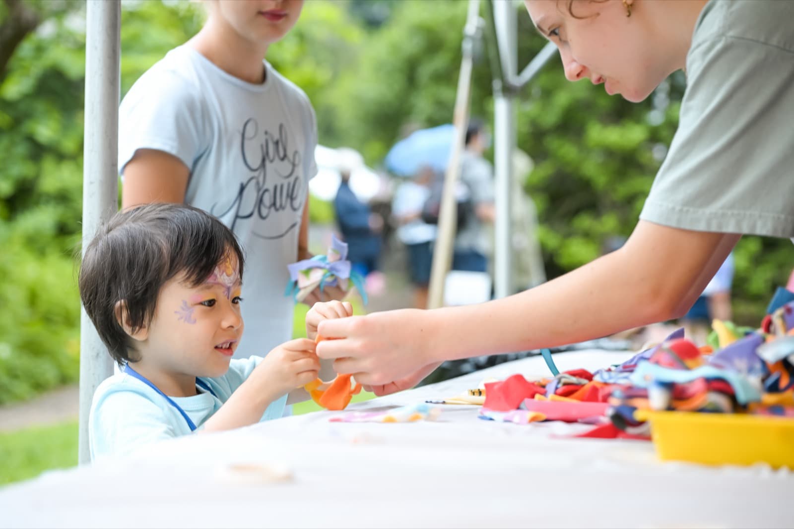 Child getting a butterfly painted on her face at a craft table, ribbons and fabric scraps spread out.