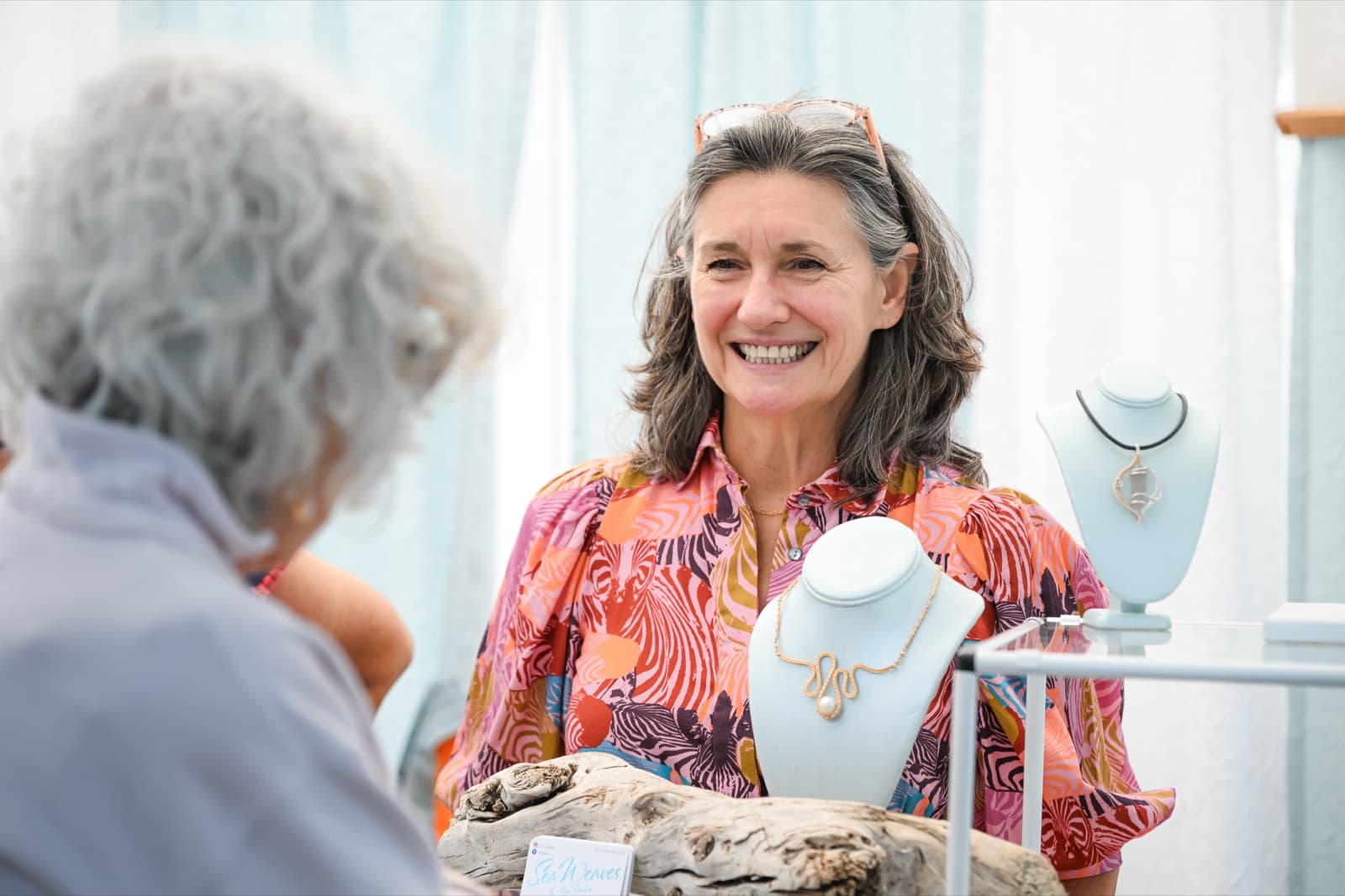 Jewelry artist smiling and chatting with a customer at her jewelry stand, driftwood and handmade necklaces on the table.