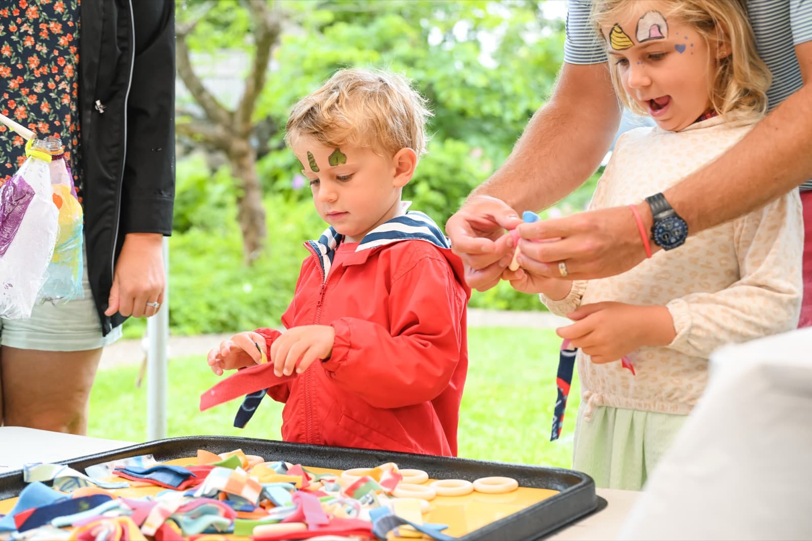 Two kids with face paint threading ribbon at a table, an adult helping guide their hands.