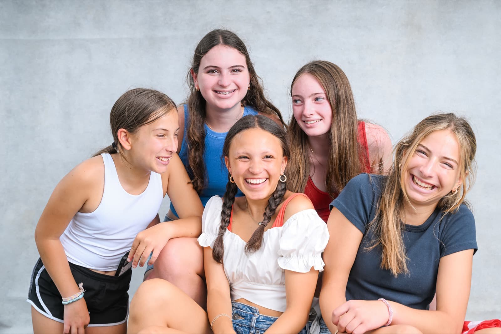 Five teenage girls laughing together in a group portrait against a plain backdrop.