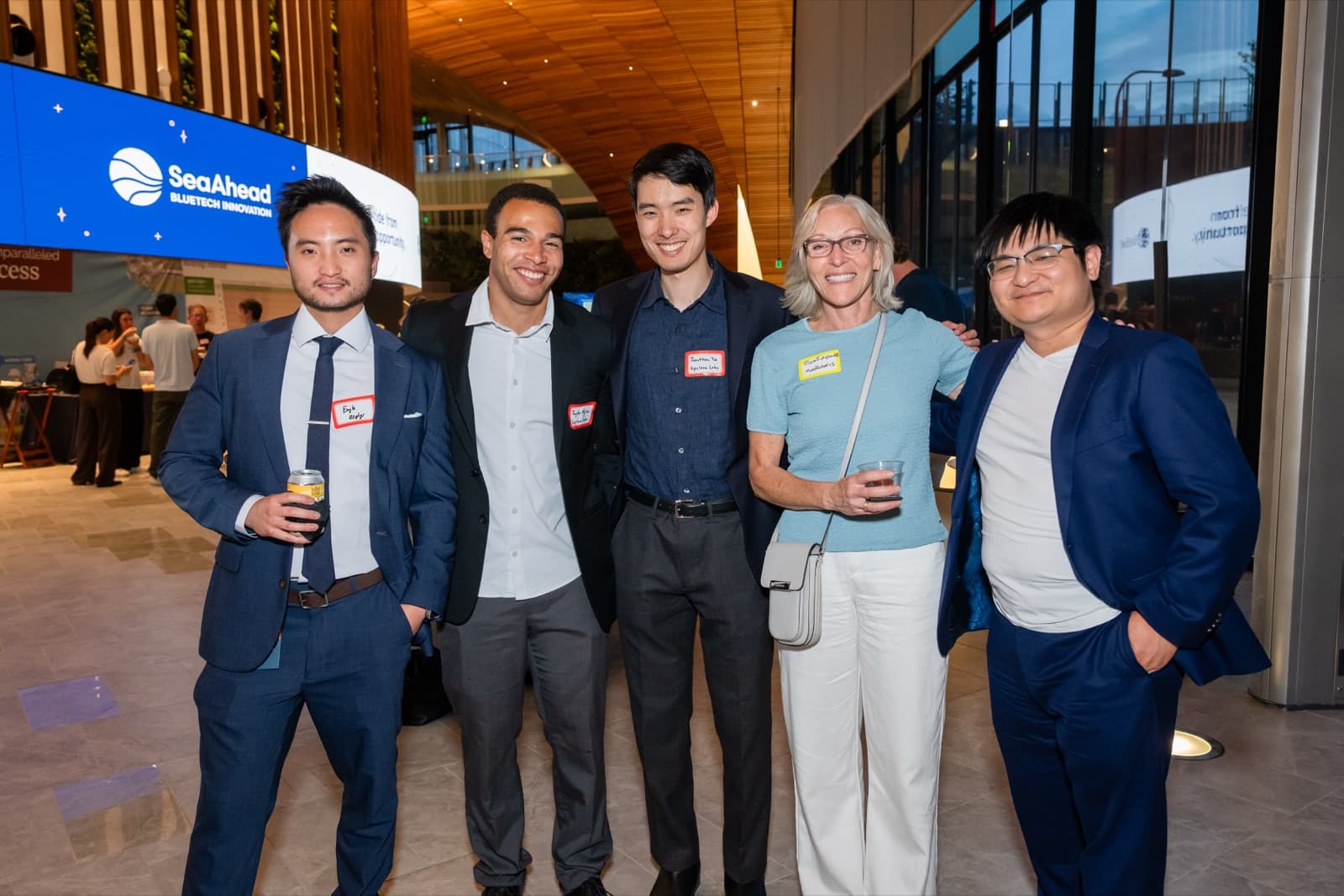 Five people posing together smiling in a modern lobby with a wood-paneled ceiling, one holding a drink.