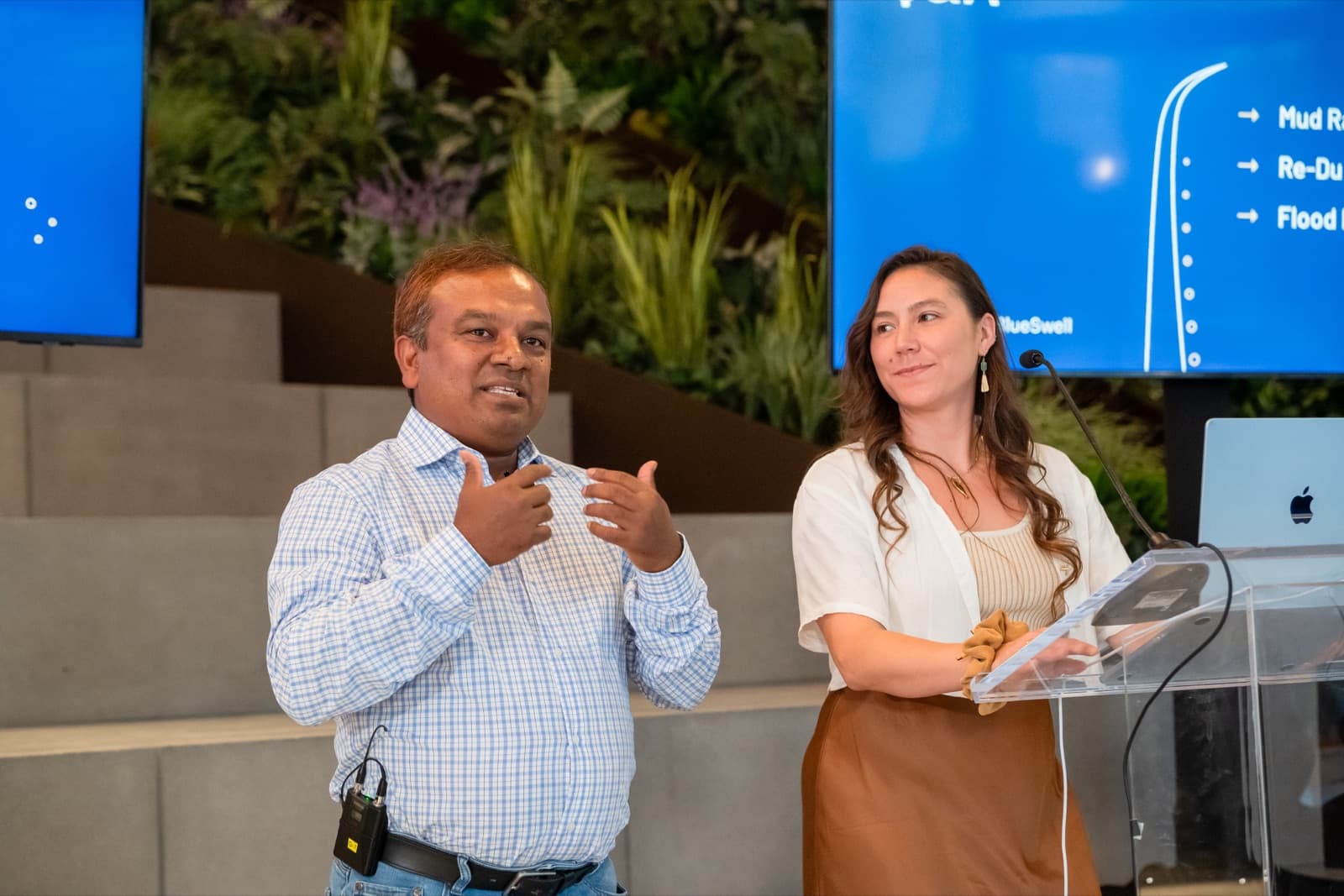 Man and woman presenting together at a clear podium, diagrams on presentation screens behind them.