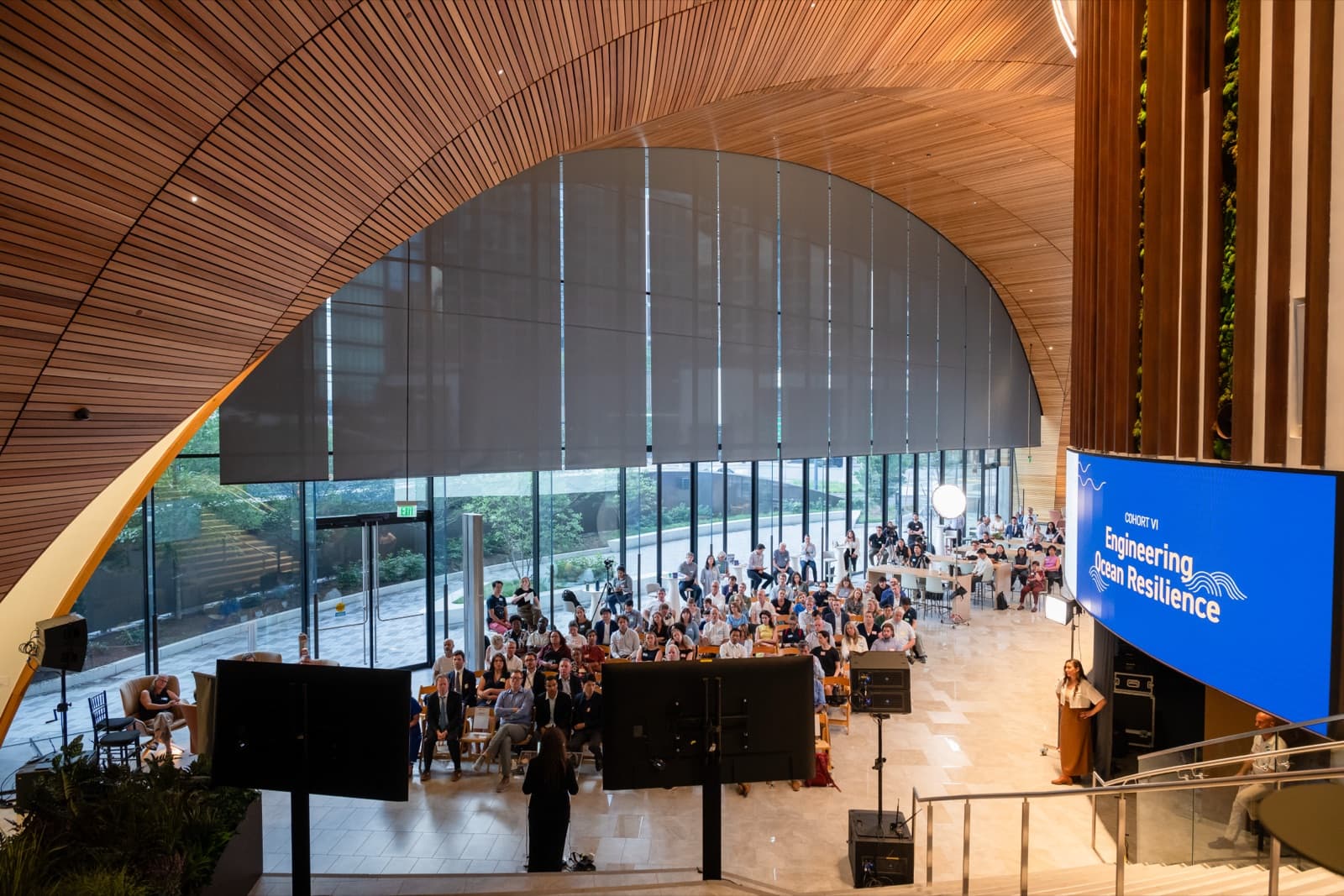 Aerial view of a large seated audience in a wood-vaulted atrium, a big screen and presenters at the front.