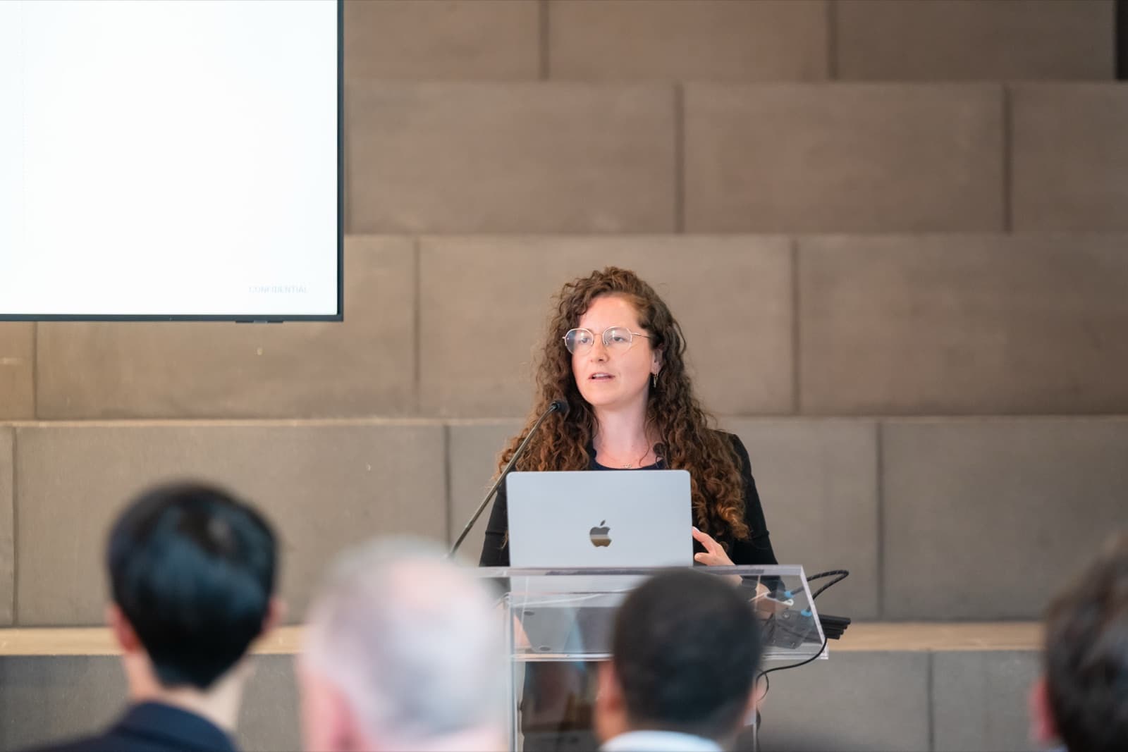 Woman with curly hair speaking at a clear podium with a laptop, a blank presentation screen behind her.
