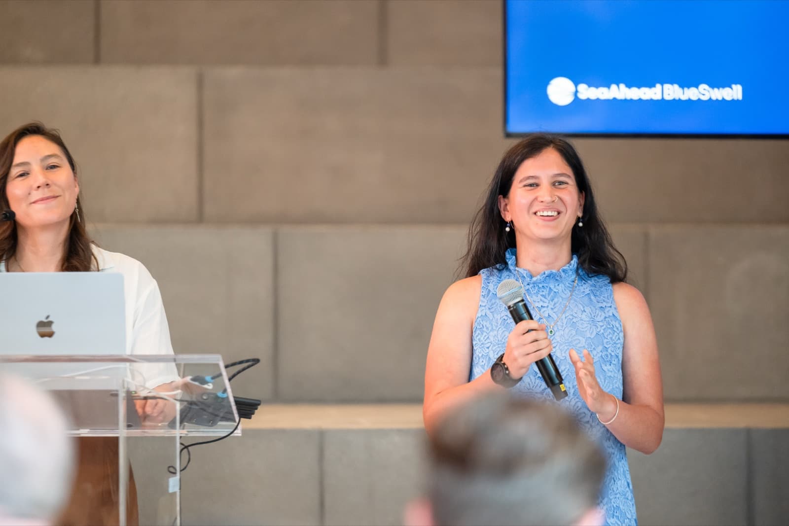 Woman in a blue lace top speaking into a microphone and gesturing, another woman standing beside a podium nearby.