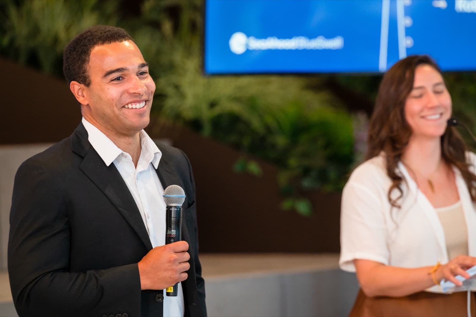 Man in a blazer speaking into a microphone, smiling, a presentation screen behind him.