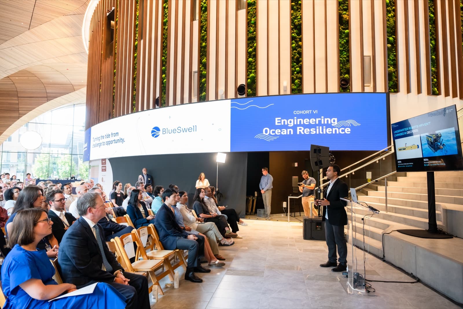 Wide view of a seated crowd listening in a wood-vaulted atrium, presenters standing near a podium at the front.