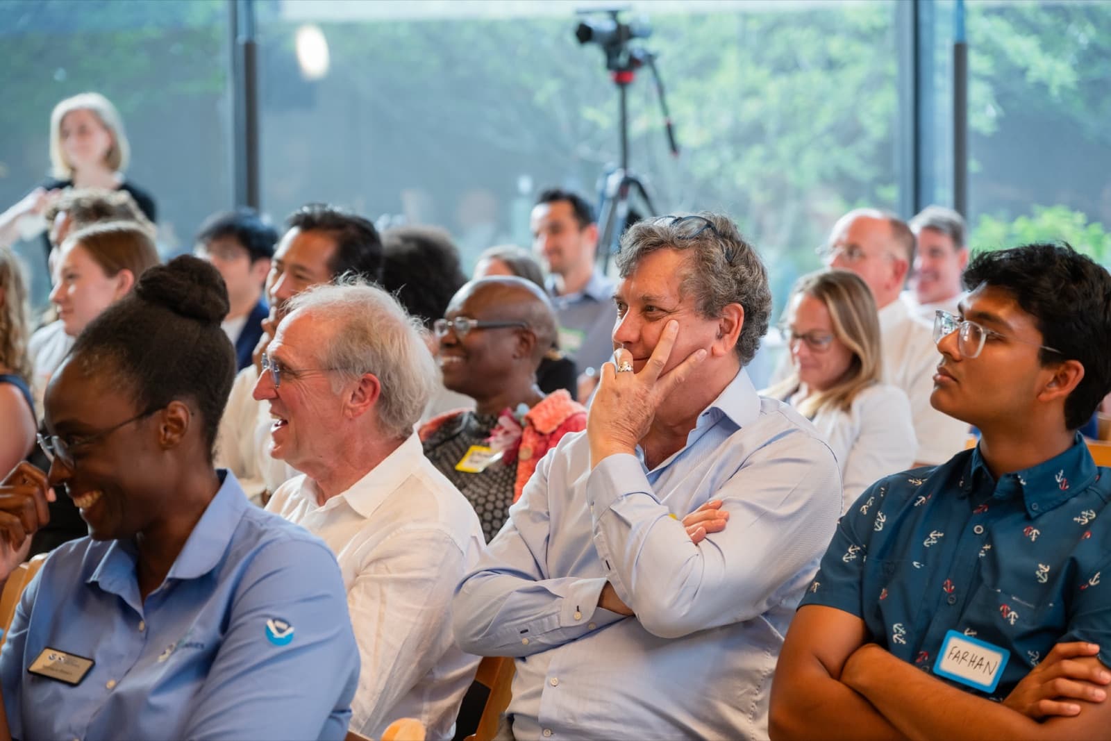 Crowd of seated attendees listening, a camera on a tripod visible in the background.