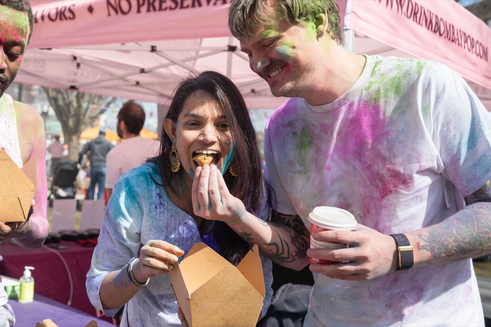 Two people covered in colorful powder, one feeding the other a bite of food from a takeout box, food tents behind them.
