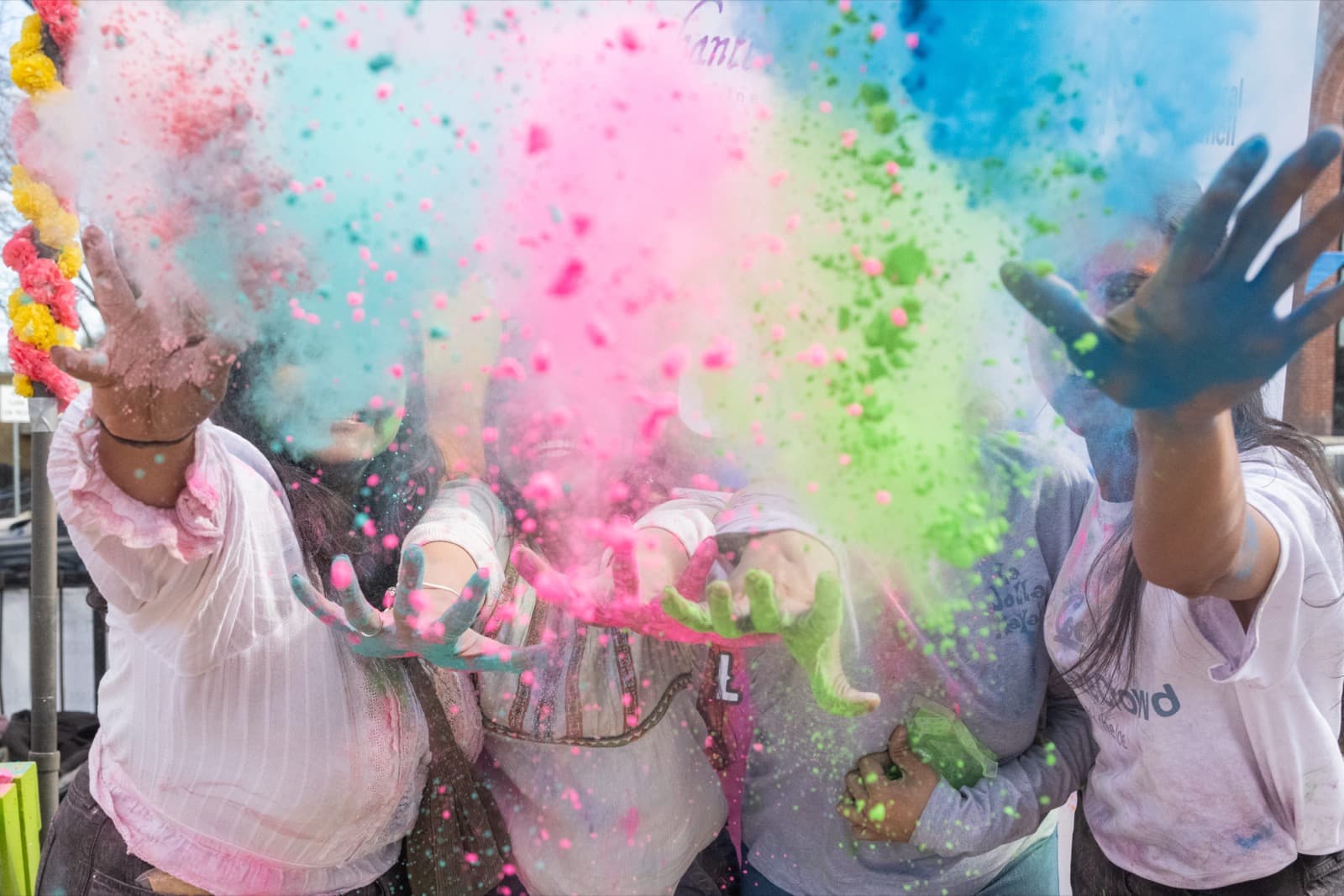 Group of people grinning as they throw handfuls of pink, teal, and green powder, clouding the air together.