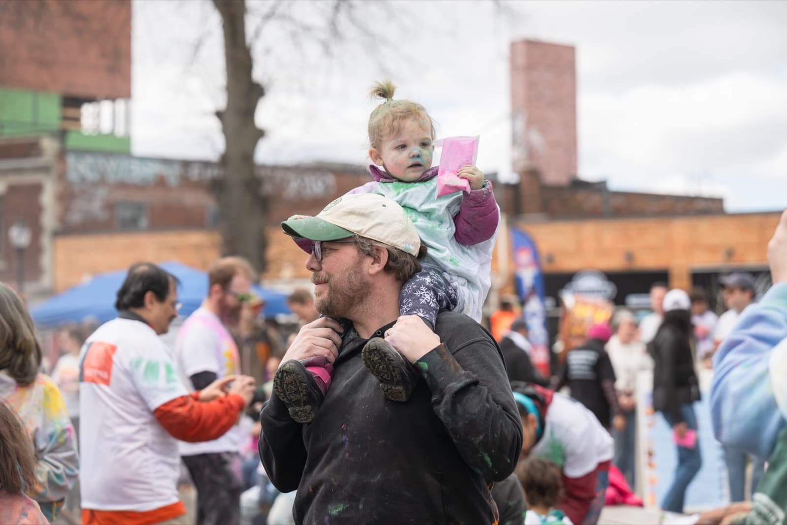 Man carrying a toddler on his shoulders through the crowd, both dusted in colored powder at a street fair.