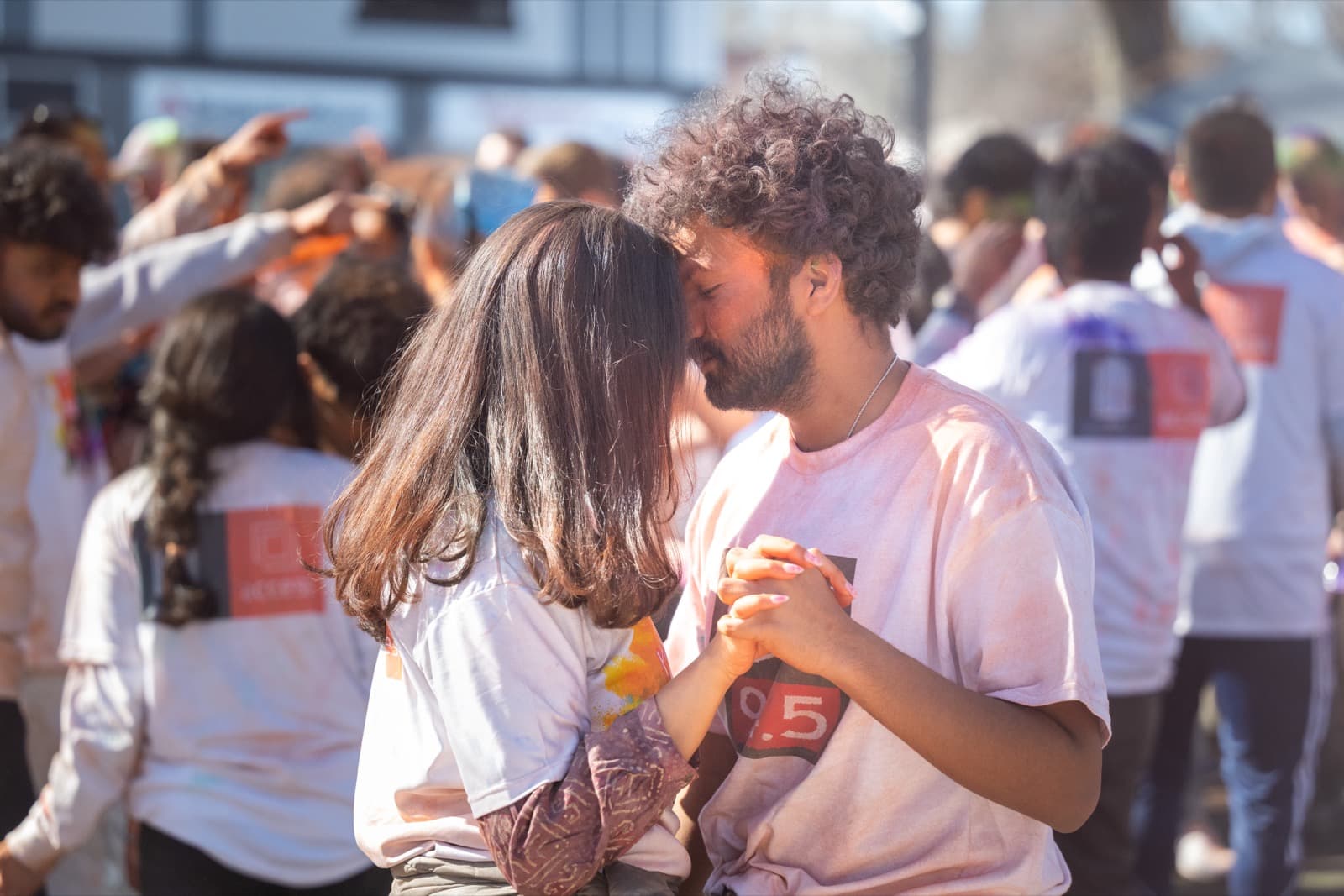 Two people with foreheads touching and hands clasped tightly between them, pink powder dusting their shirts.