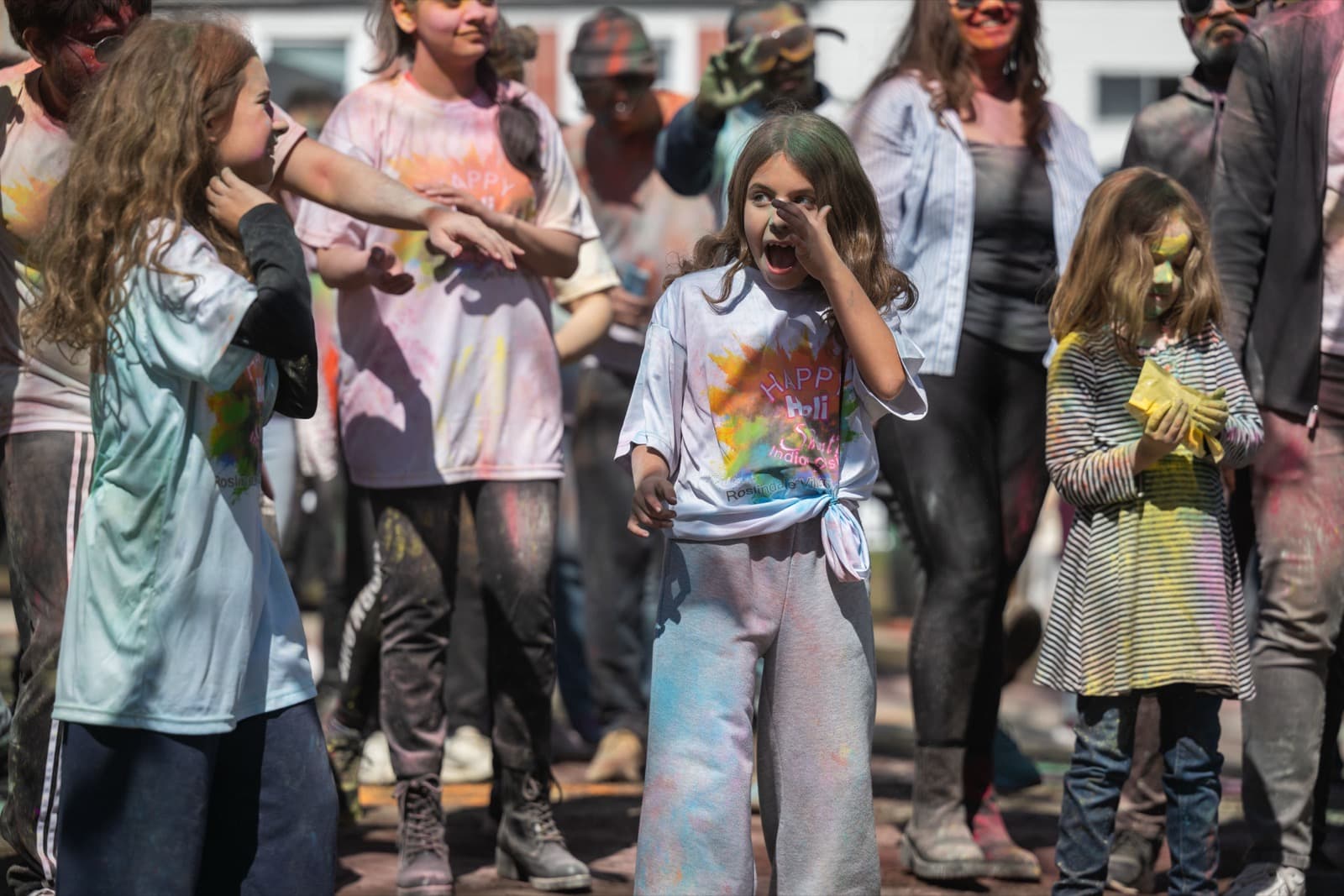 Girl in a “Happy Holi” t-shirt gasping in delighted surprise, colorful powder streaked across her shirt.