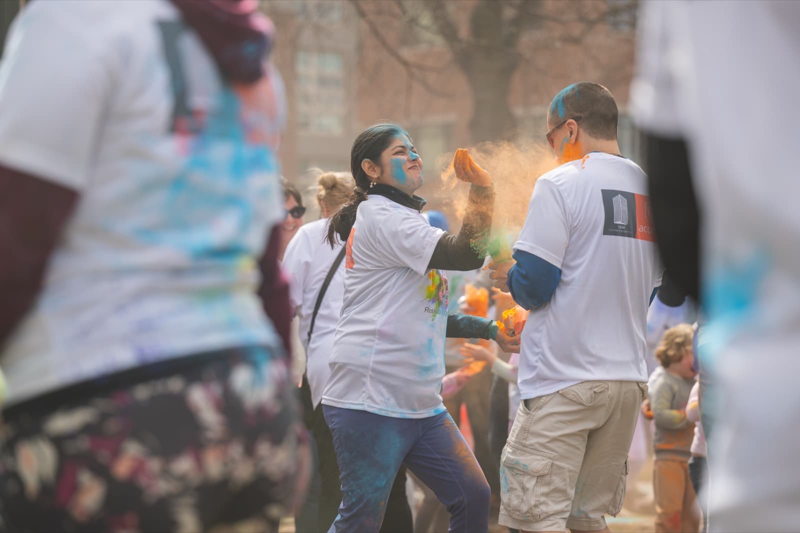 Two people in matching event t-shirts, laughing as they throw handfuls of orange powder at each other.