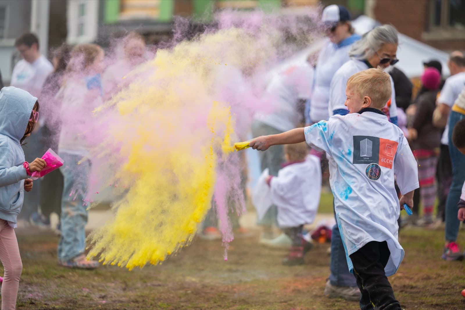 Young boy in a branded event shirt flinging a burst of yellow powder overhead, the cloud filling the frame.
