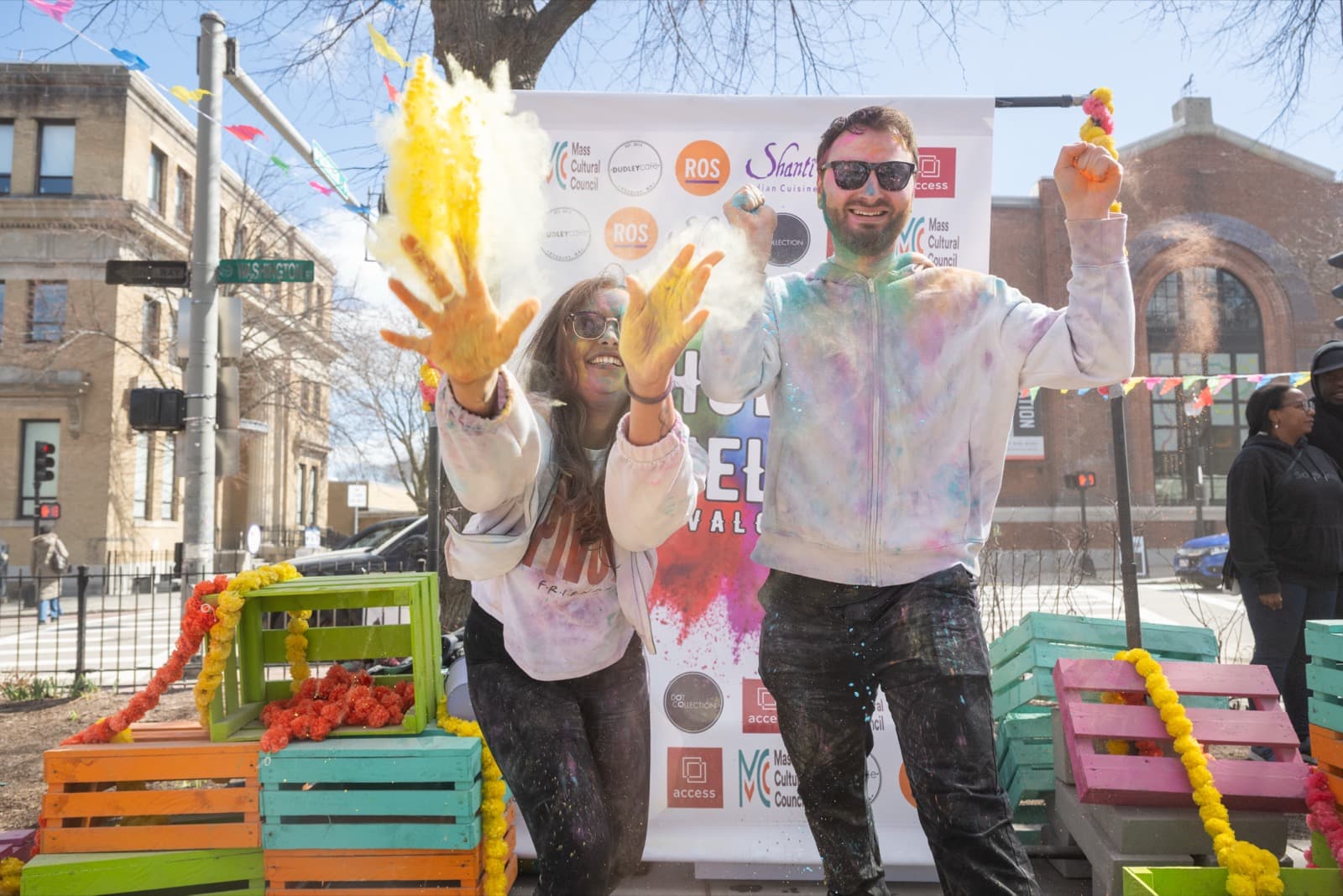 Two people celebrating in front of a festival banner, throwing yellow powder above marigold-strung crates.