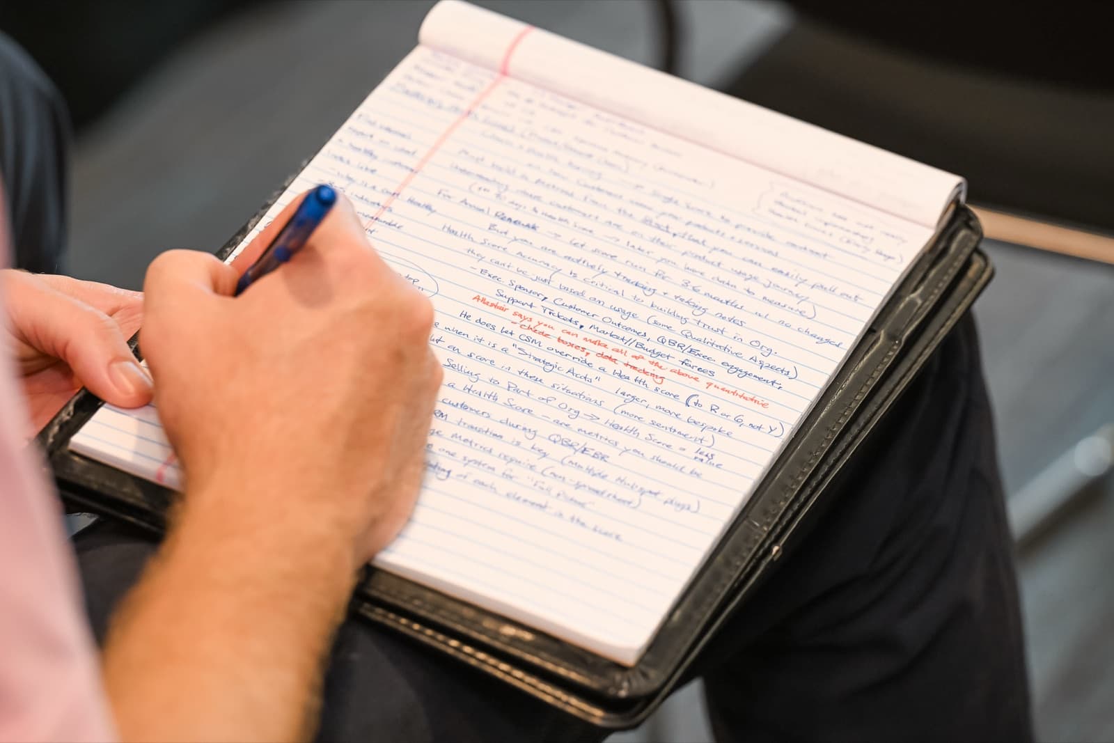 Close-up of a hand writing detailed notes with a pen in a spiral notebook, resting on a lap during a session.