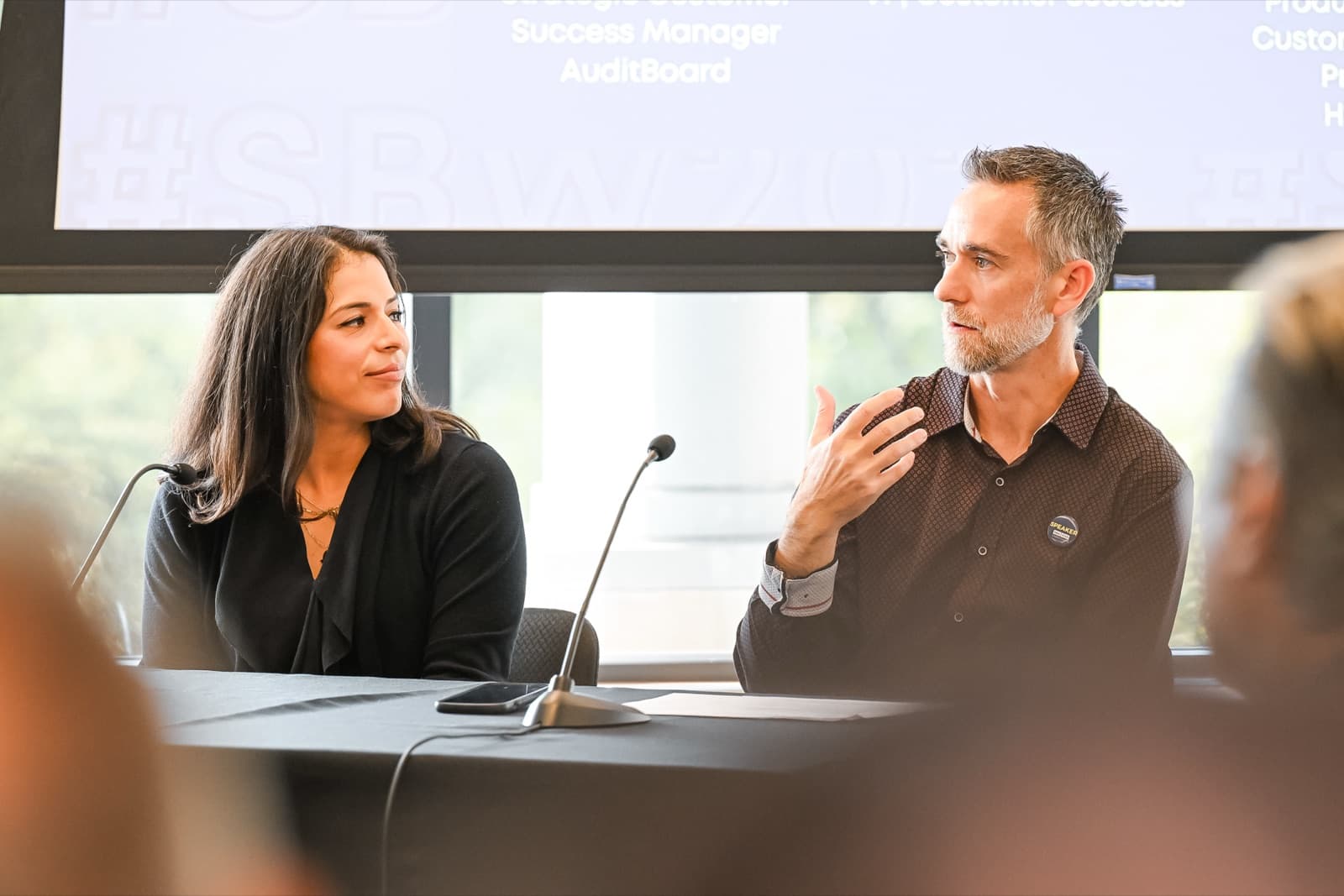 Man and woman seated on a panel with lapel microphones, a presentation screen behind them.