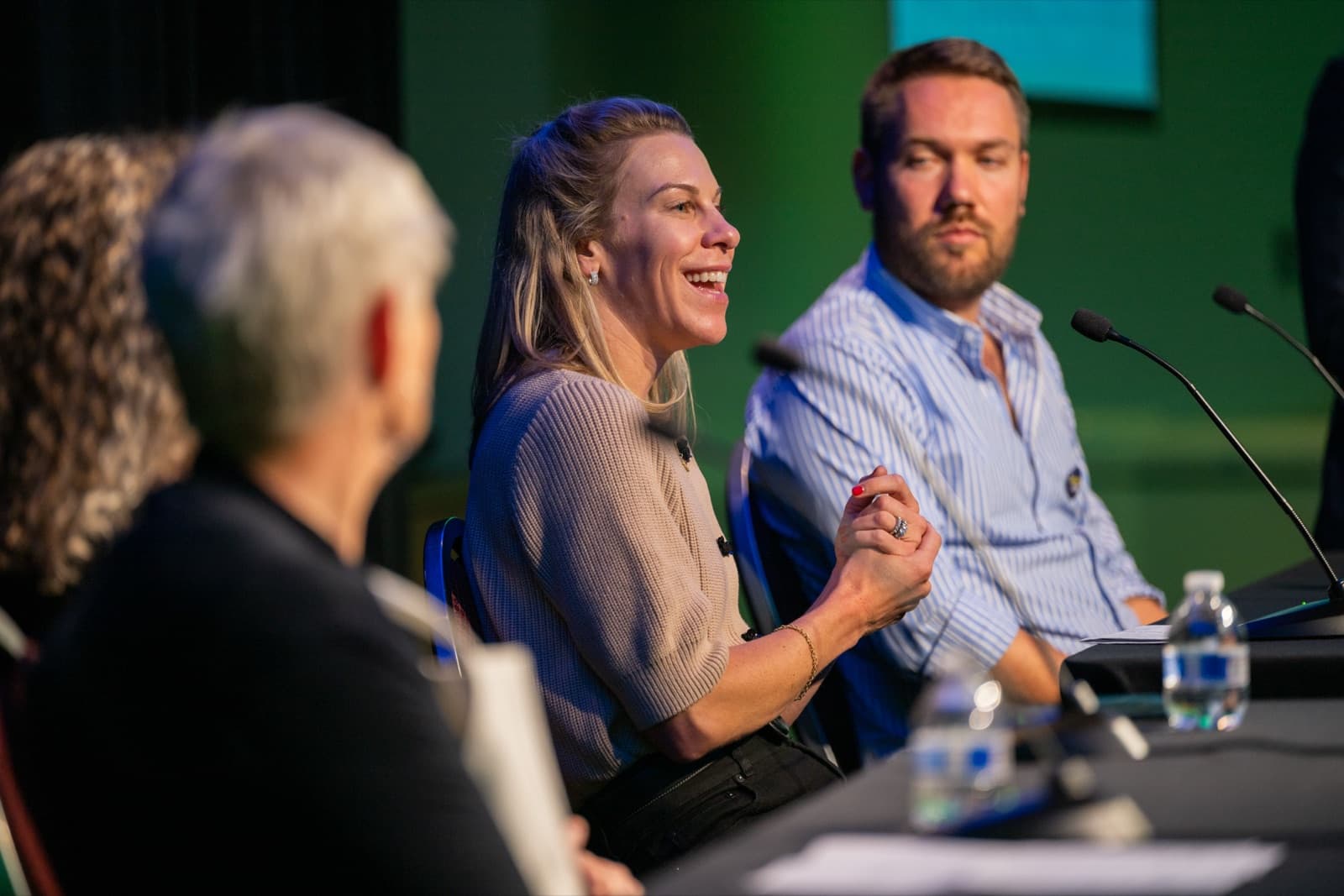 Woman grinning and gesturing while seated on a panel, two other panelists beside her, a green-lit stage.
