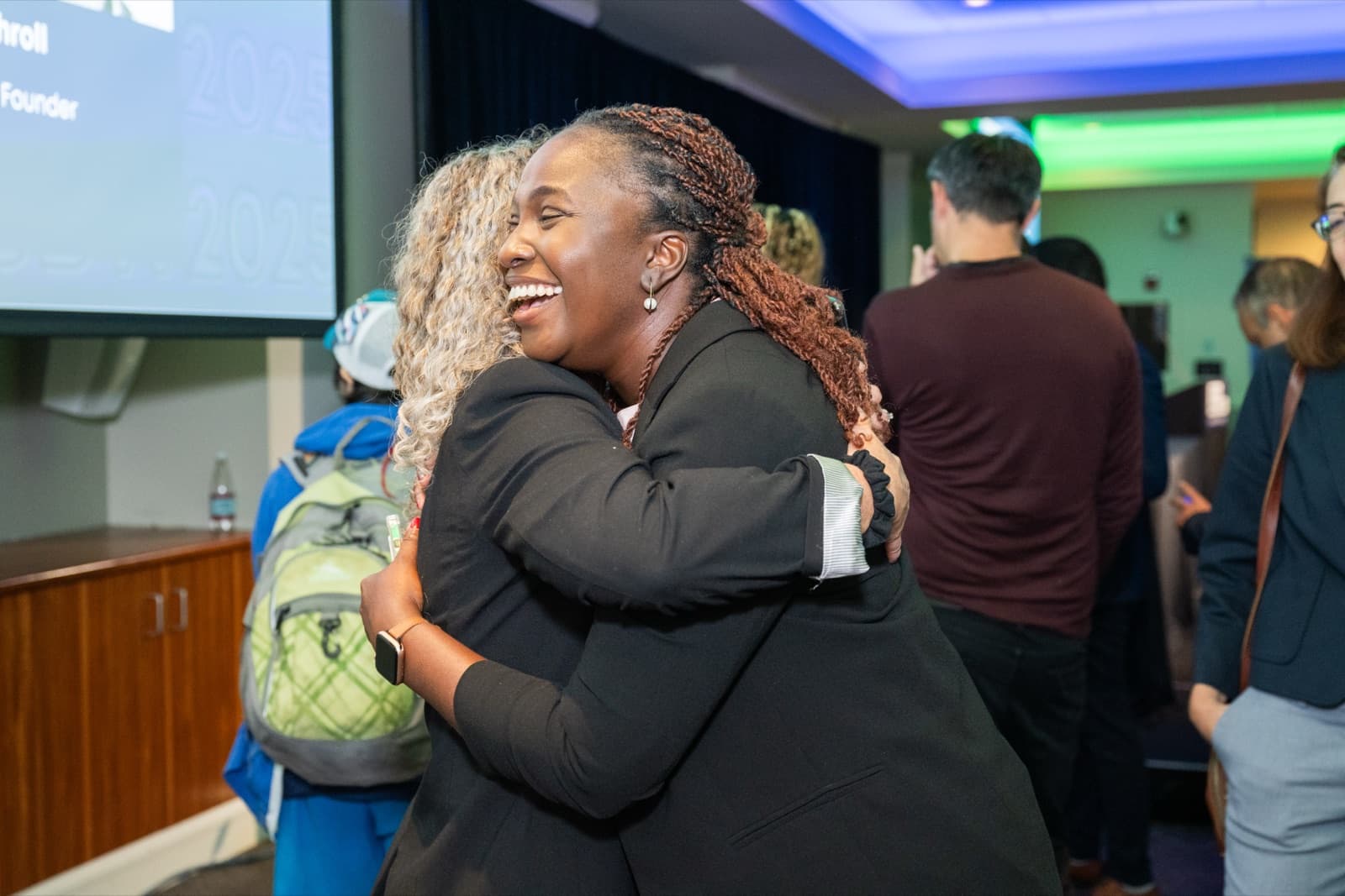 Two women embracing and laughing warmly, other attendees visible behind them.