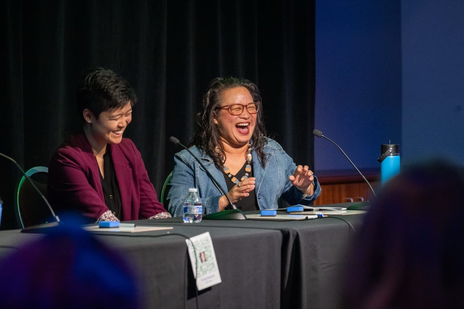 Two women laughing together on a panel, seated behind a table with microphones.