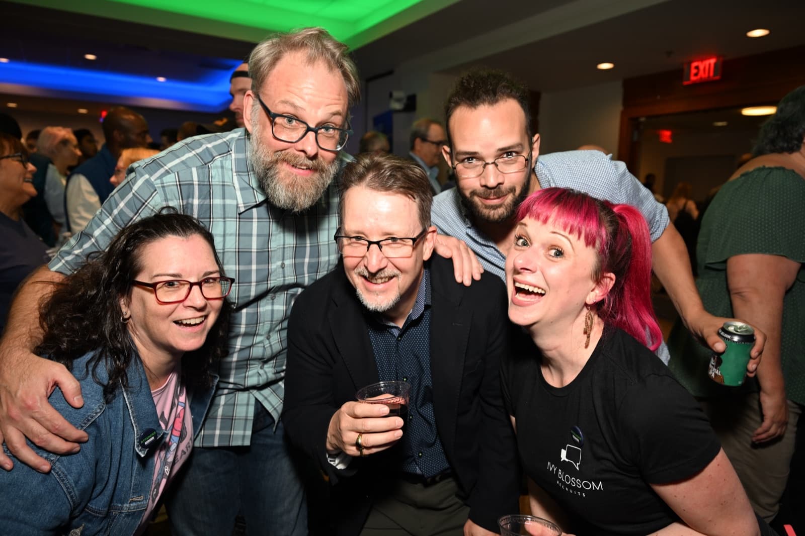 Five people smiling together, one holding a drink, in a crowded reception.