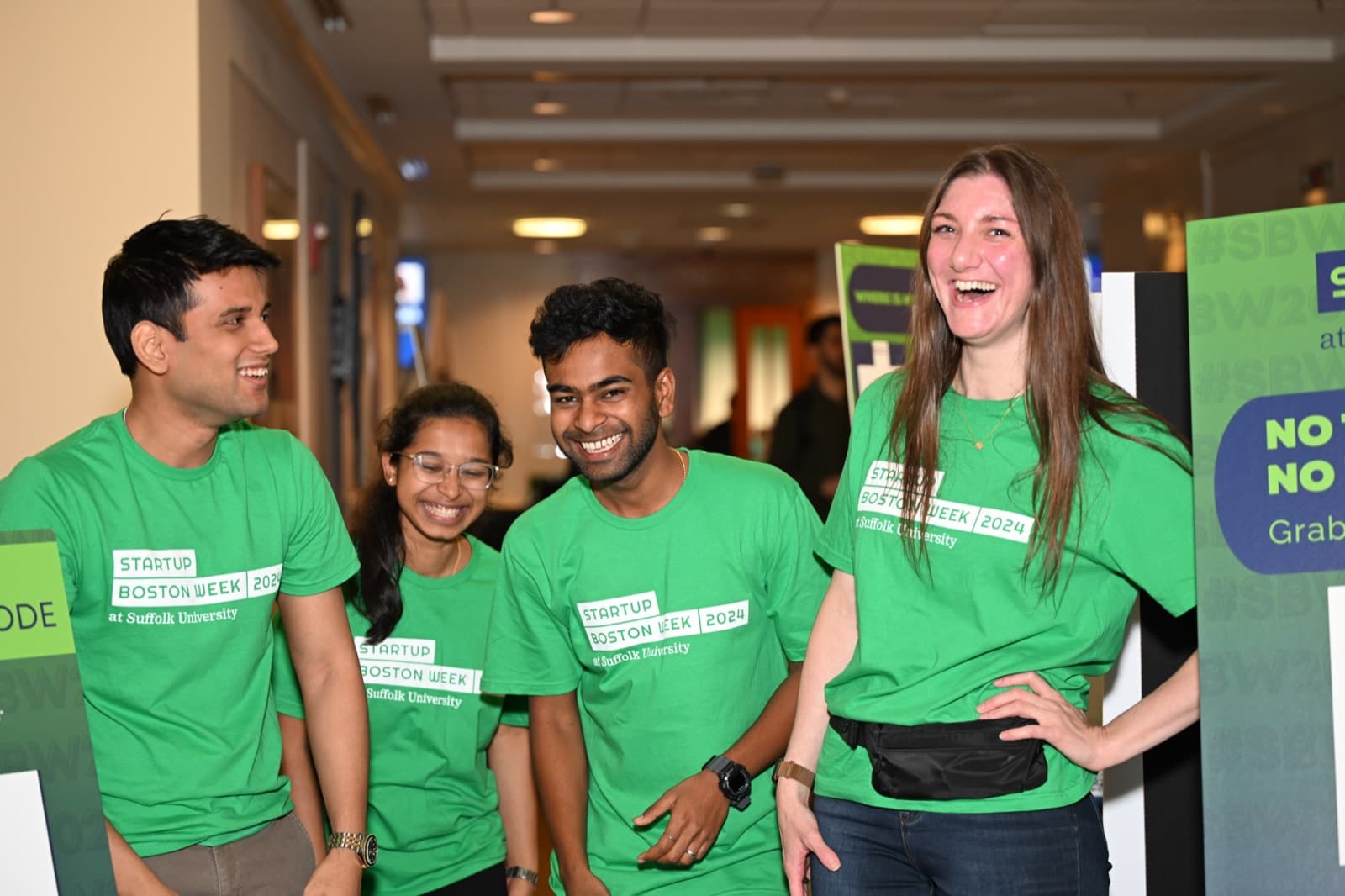 Four people in matching green event t-shirts smiling together in a hallway, event signage visible nearby.