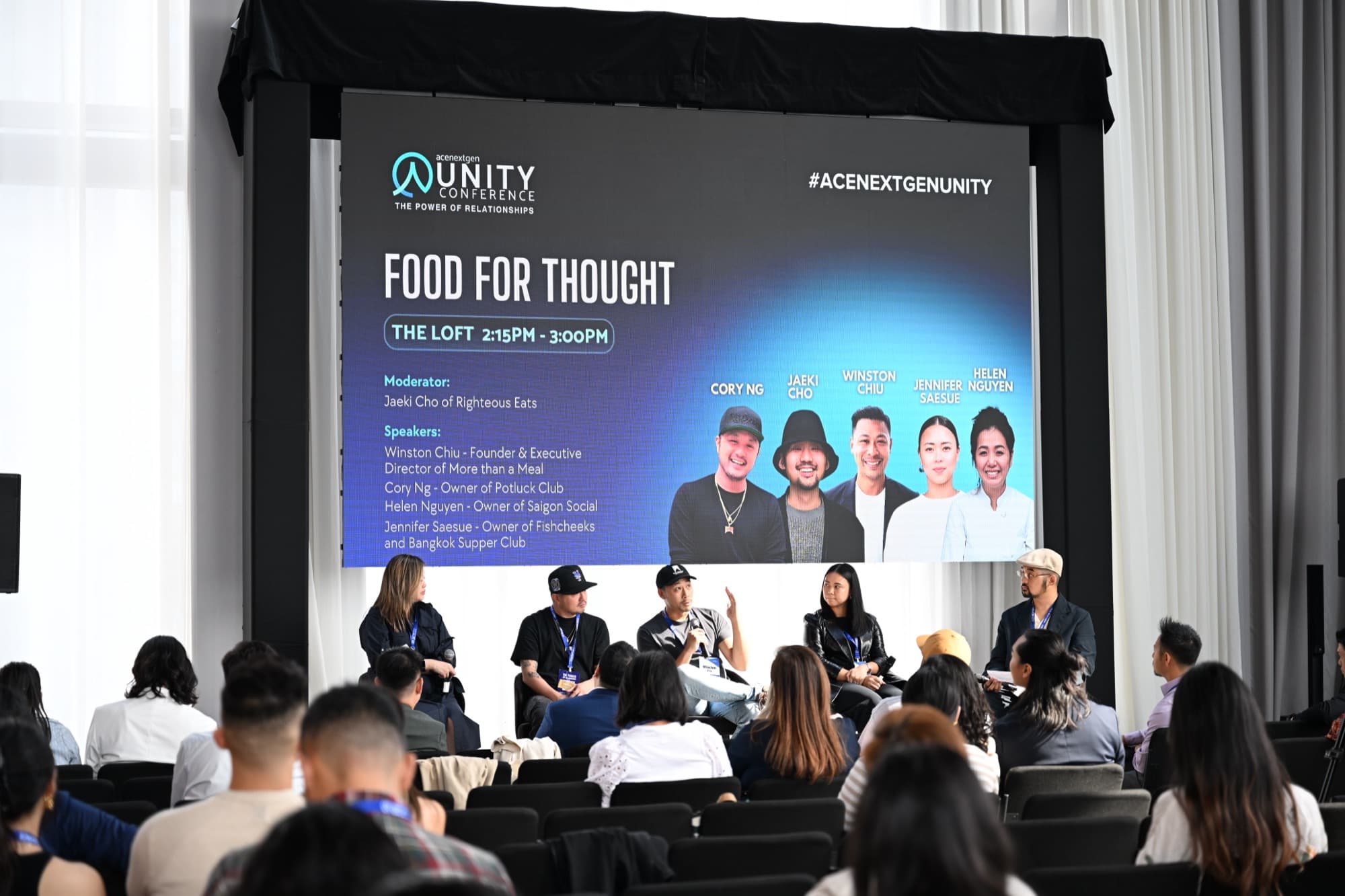 Five panelists seated on stage for a discussion, a large screen behind them, an audience in front.