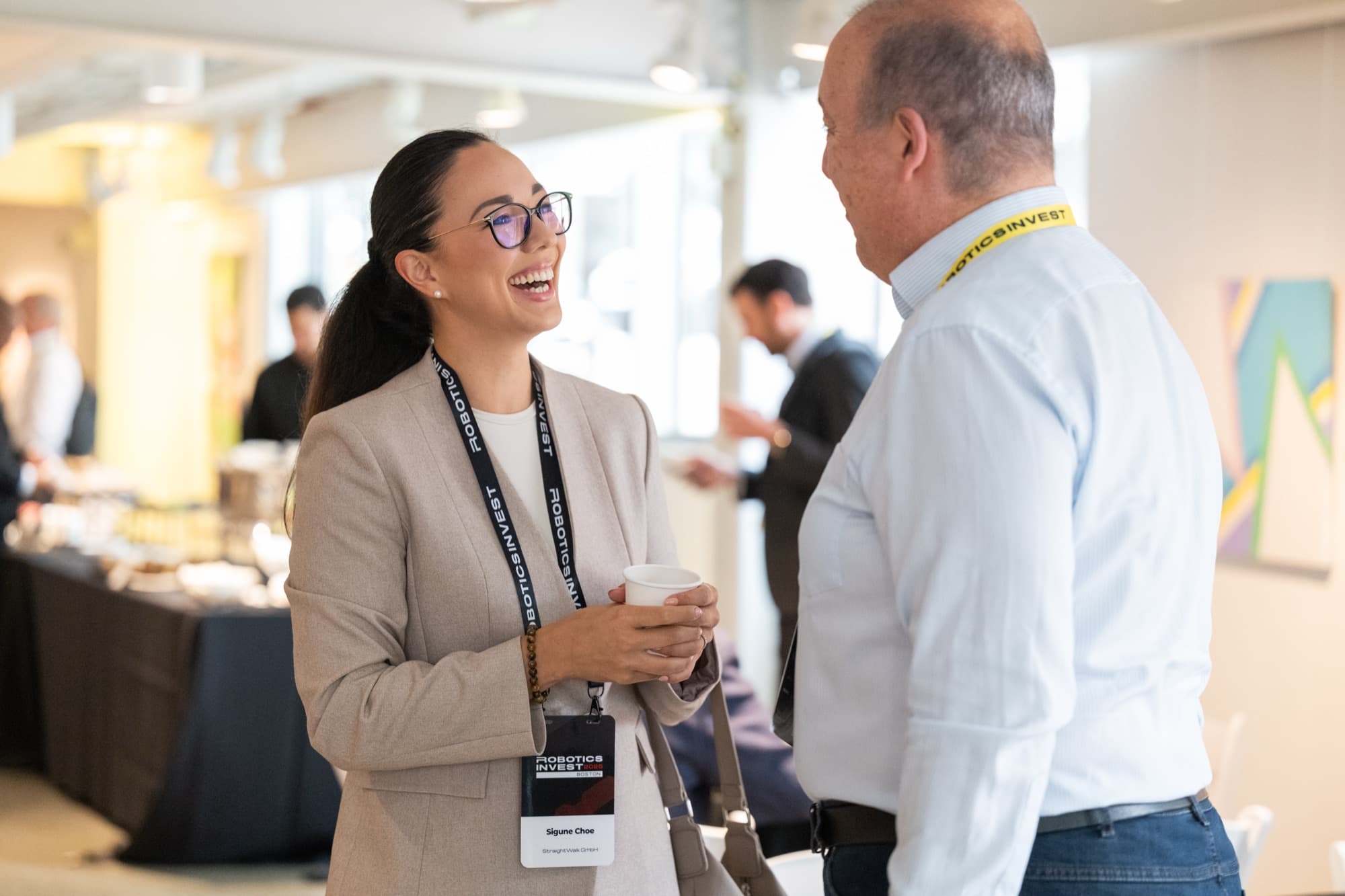 Woman laughing while holding a coffee cup, talking with a man at a conference networking break.