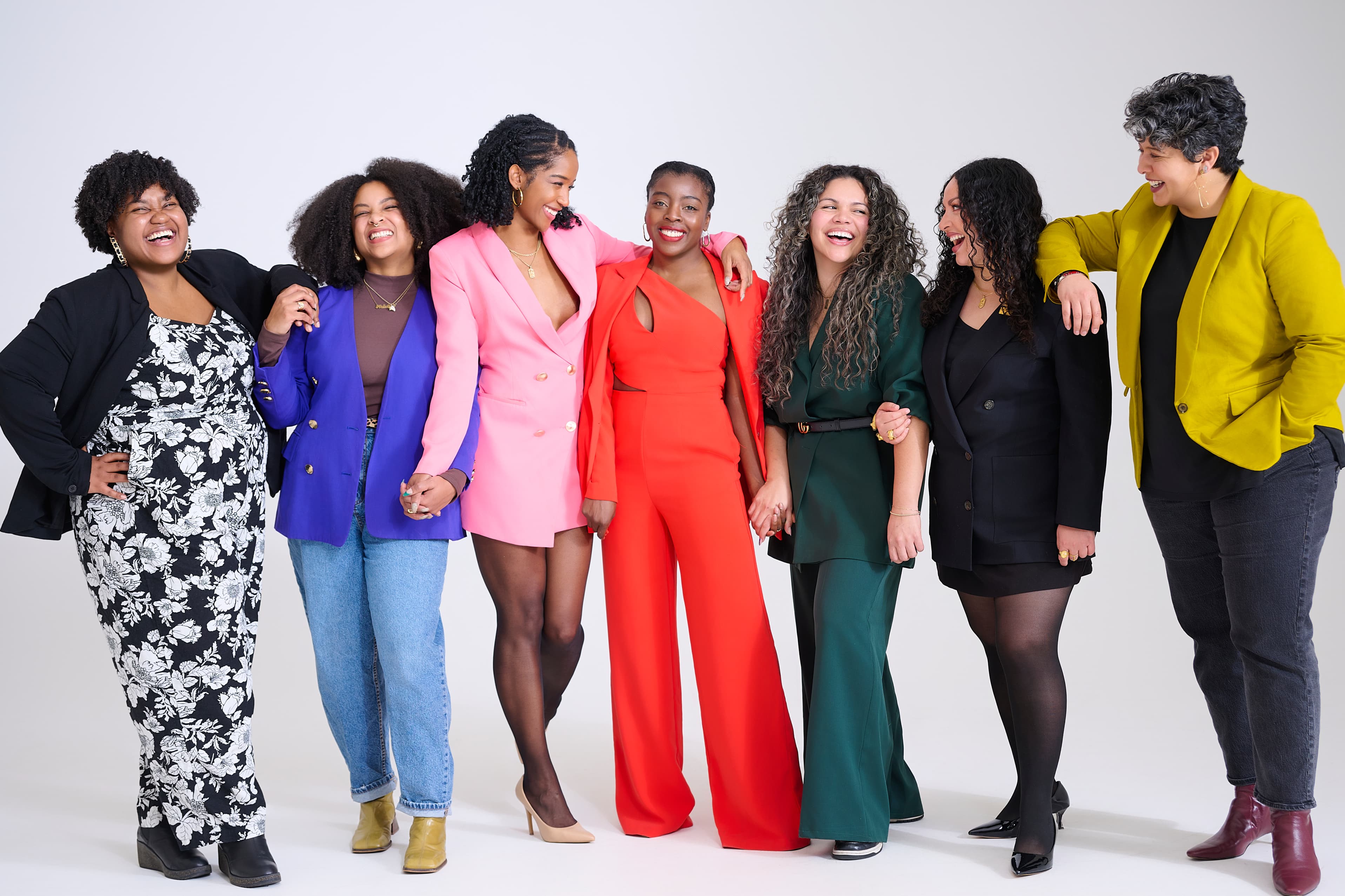 Seven women in colorful blazers and dresses laughing together, arms linked, in a bright studio.