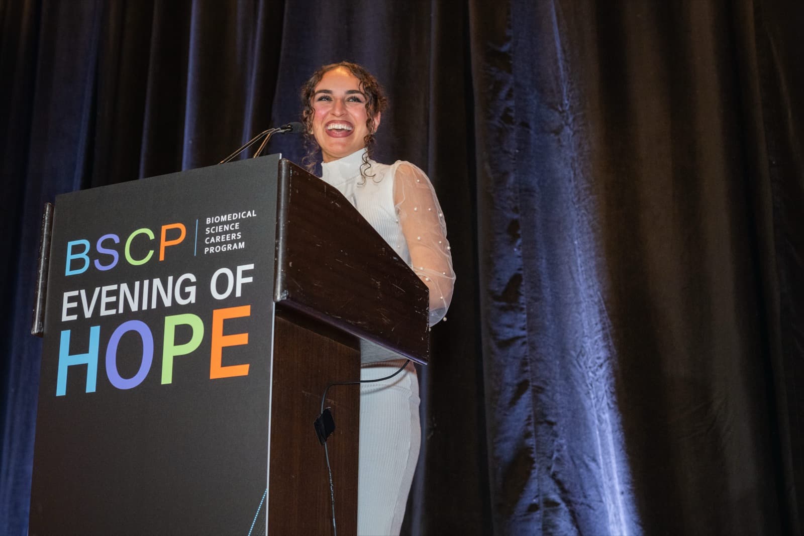 Woman in a sheer pearl-sleeved top laughing at a podium, a dark curtain behind her.