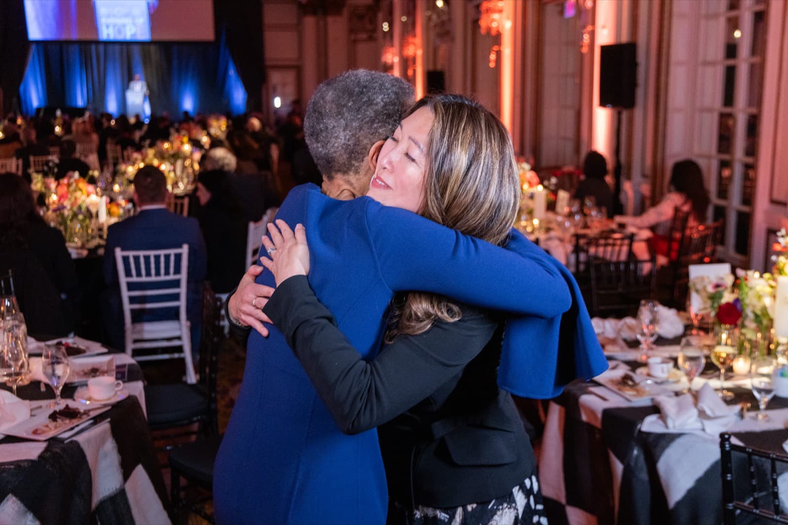 Two women embracing warmly, one in a royal blue dress, a grand chandelier-lit room behind them.