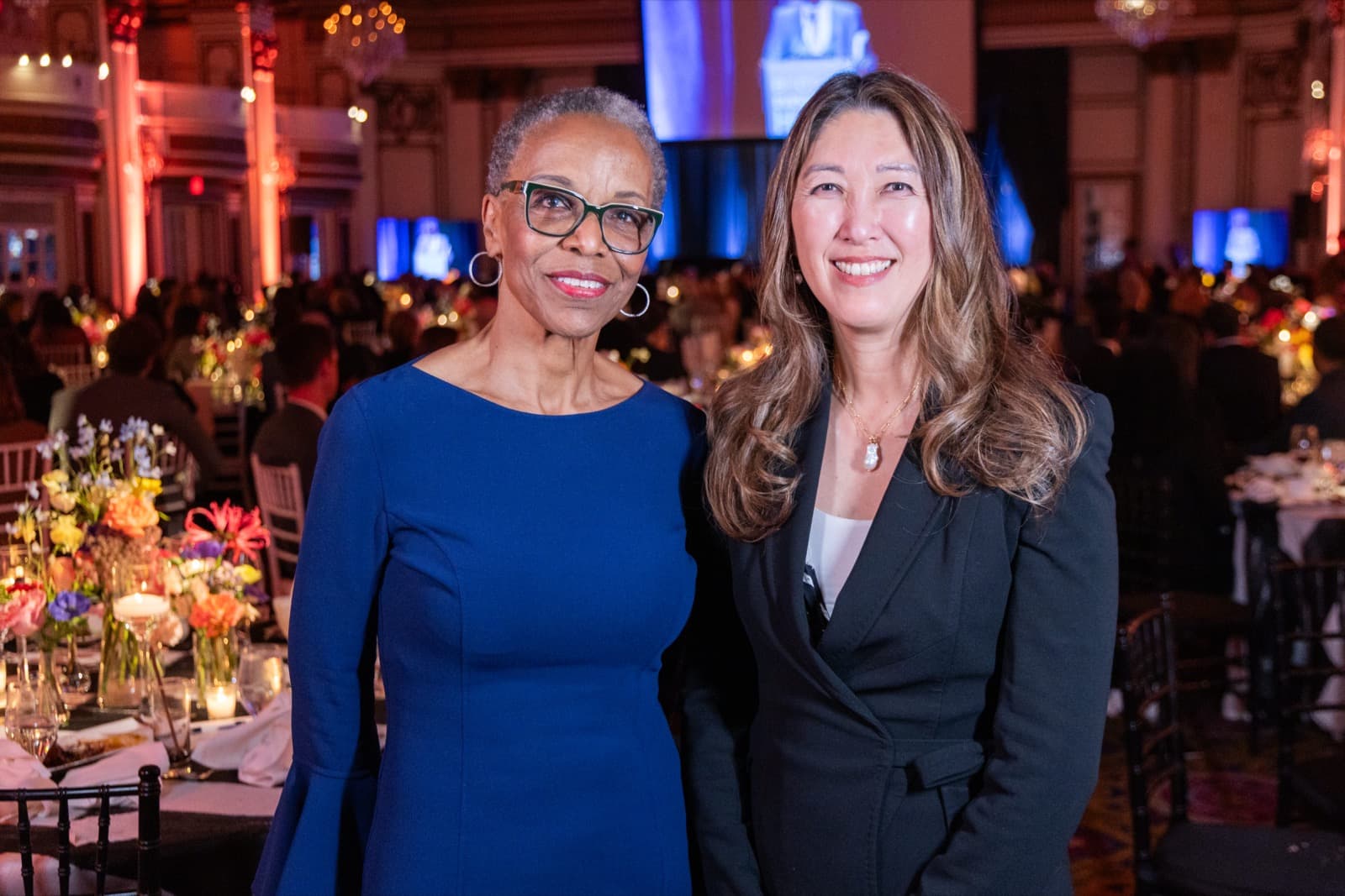 Two women posing together smiling, one in a blue dress and glasses, an ornate hall behind them.