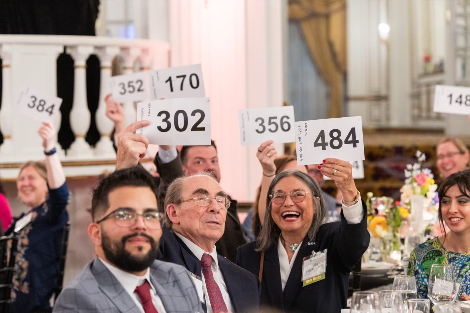 Gala guests holding up numbered paddles at their table, floral centerpieces and an ornate balcony railing nearby.