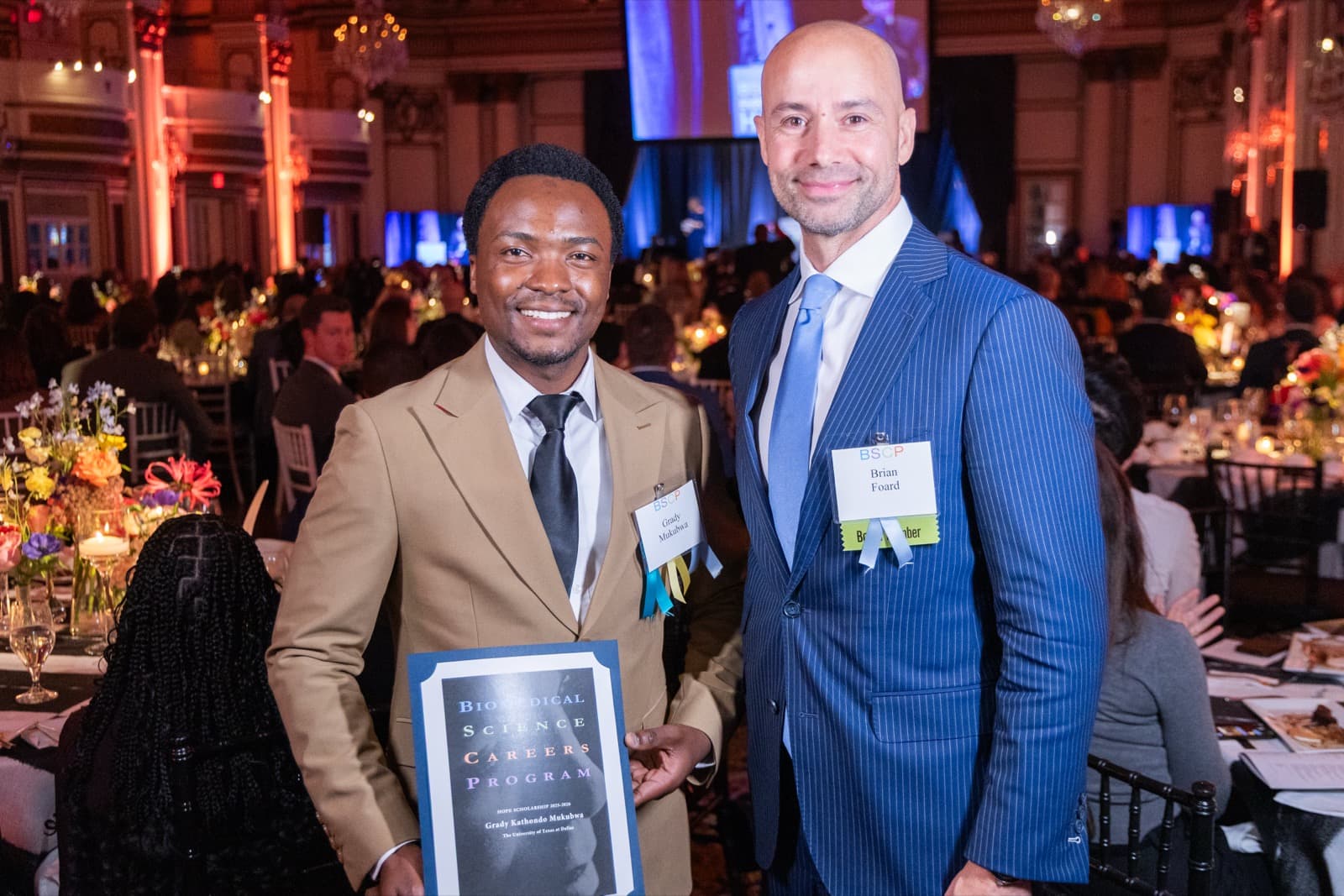Two men smiling together, one in a tan blazer holding a certificate, a crowded reception behind them.