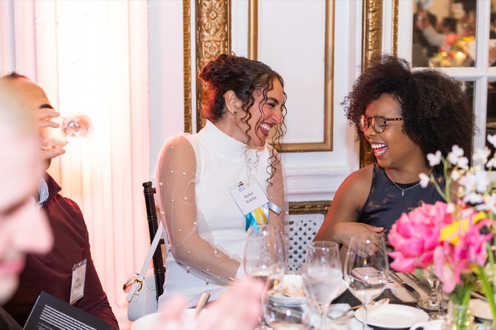 Two women laughing together at a dinner table, one in sheer pearl-beaded sleeves, gilded paneling behind them.