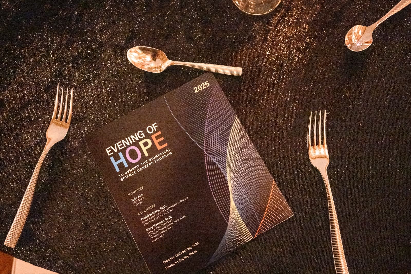 Printed event program on a dark tablecloth, listing an honoree, co-chairs, and a date, silverware nearby.