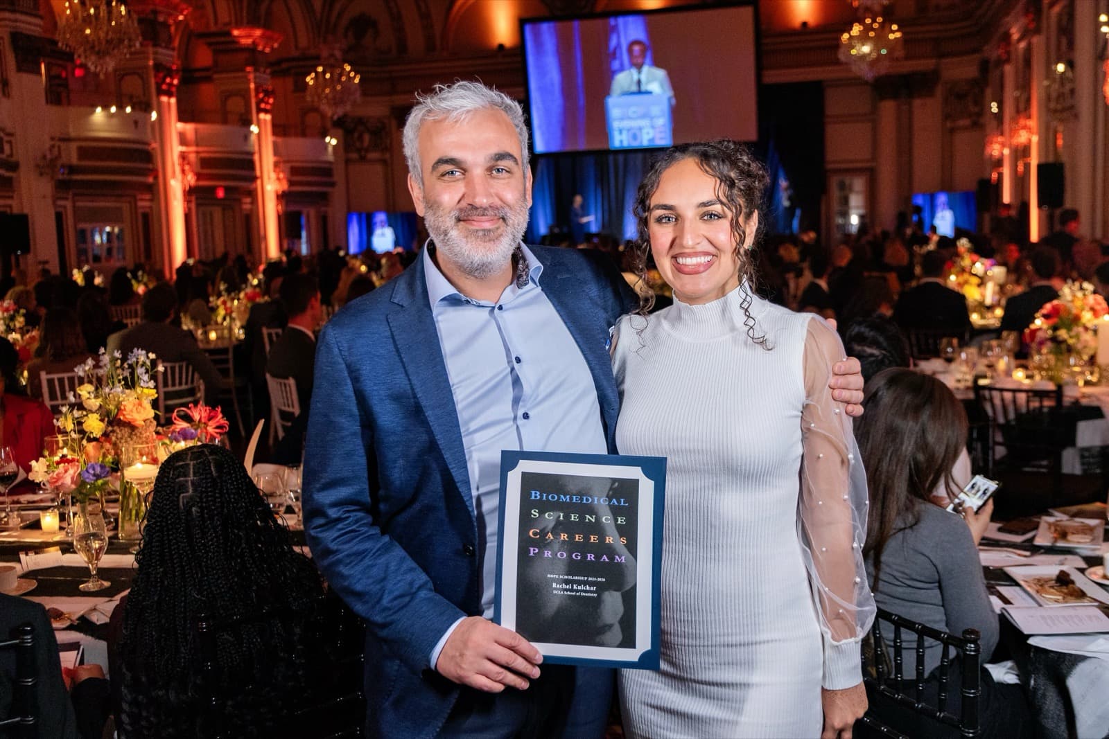 Man with gray hair and a beard holding a certificate, arm around a smiling woman in a white dress, a crowded hall behind them.