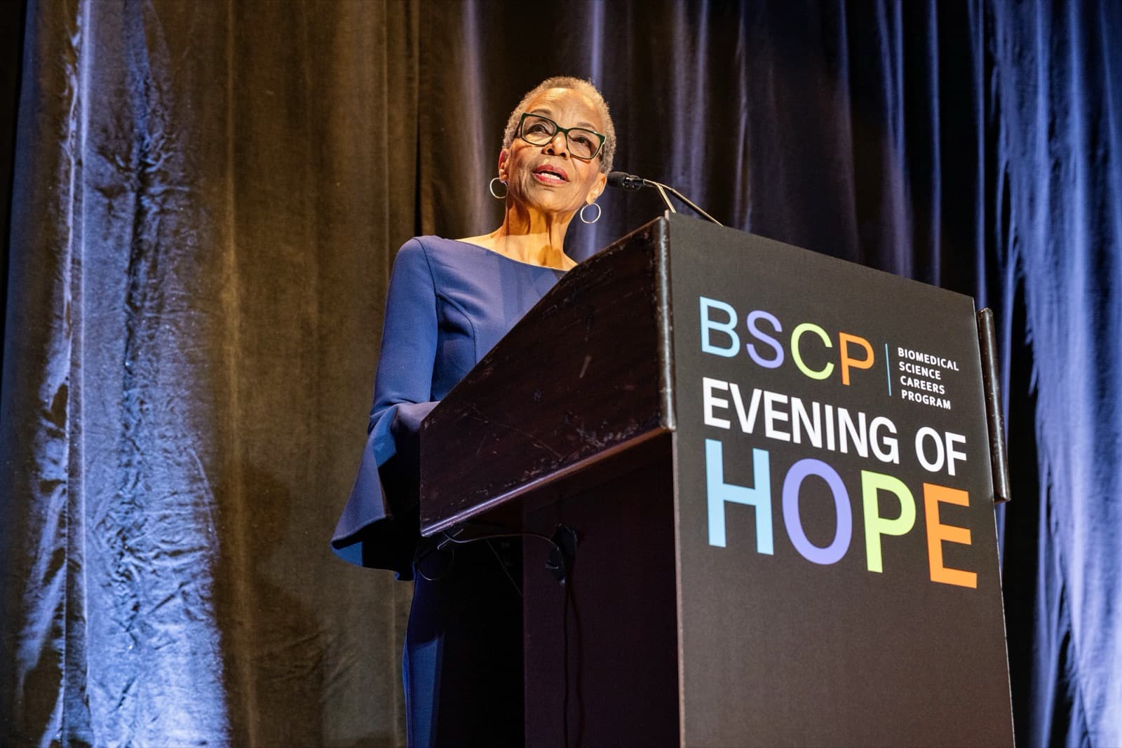 Woman with short gray hair and glasses speaking at a podium, a dark curtain behind her.