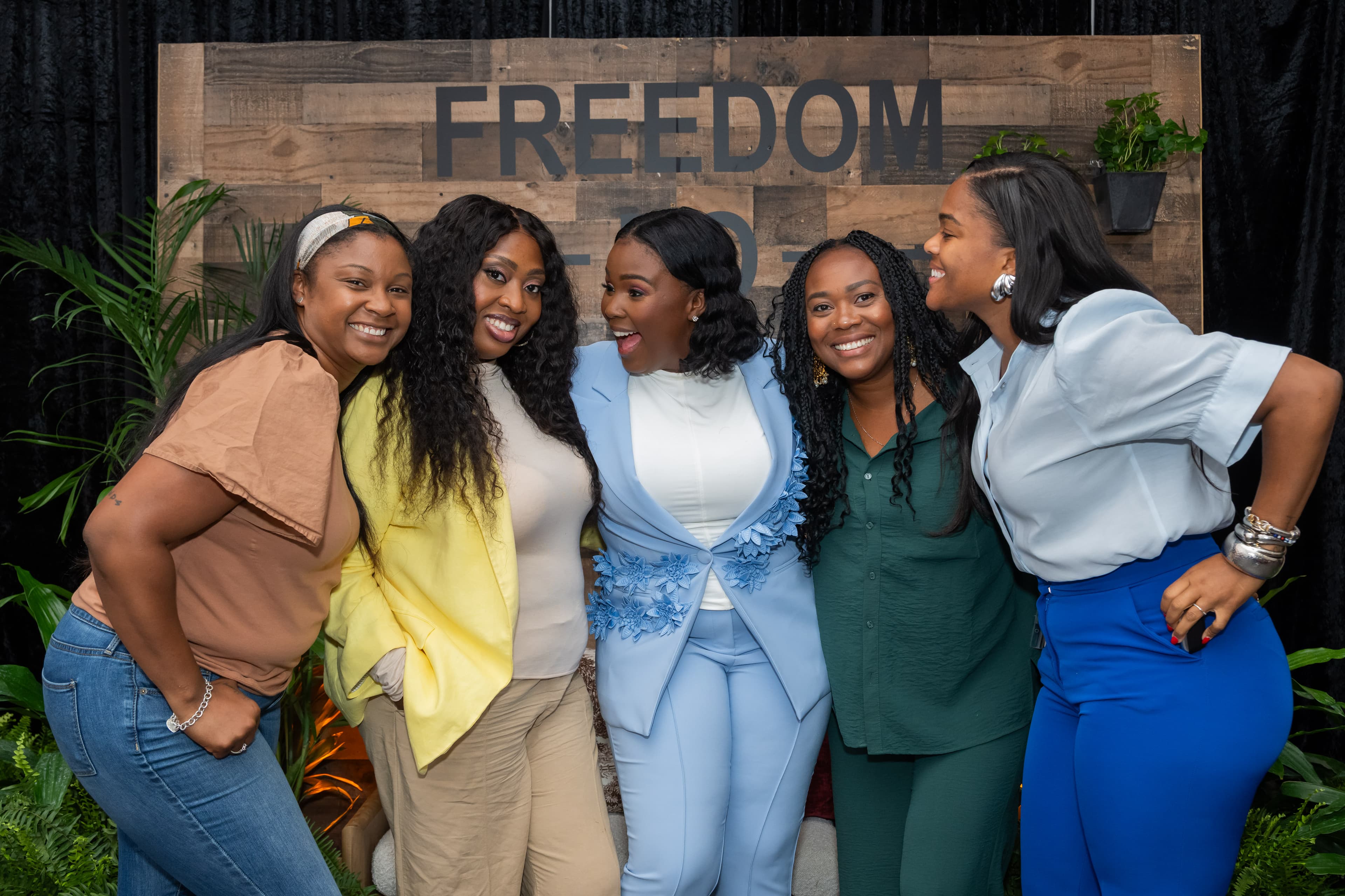 Five women laughing together in front of a wooden accent wall, tropical plants nearby.