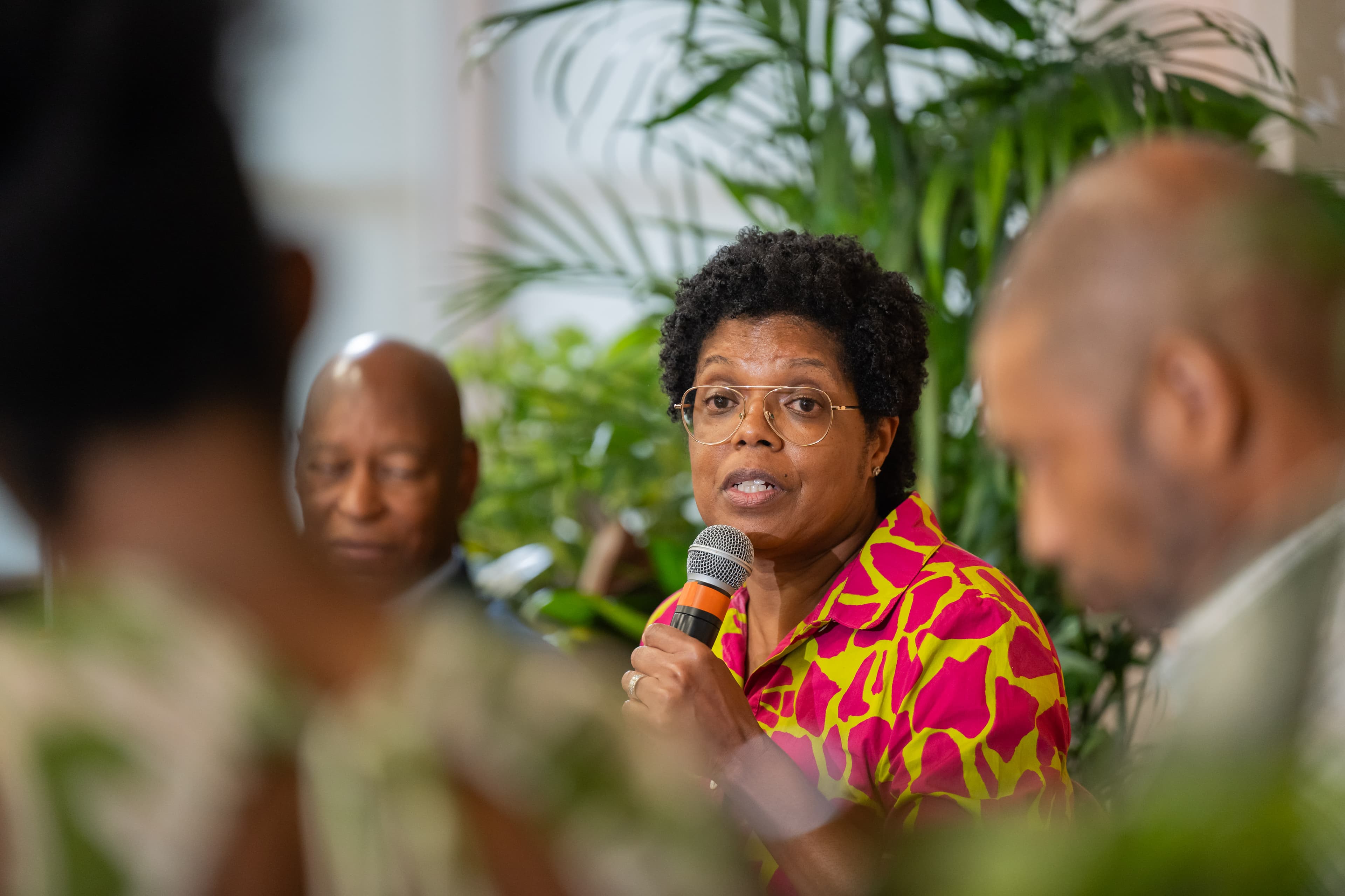 Woman gesturing while speaking into a microphone, two men blurred beside her, tropical plants behind her.