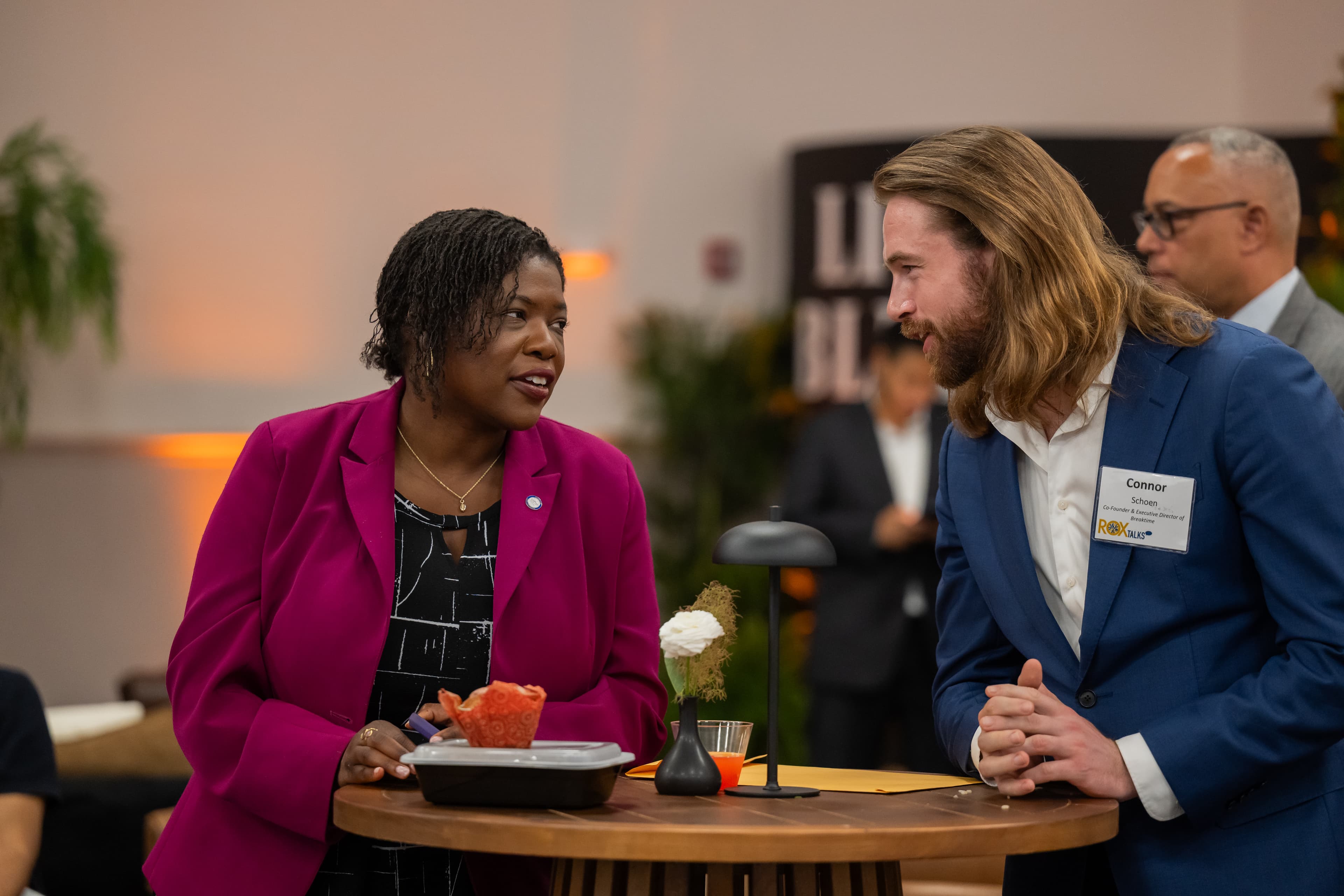 Woman in a magenta blazer talking with a man with long hair and a beard, both smiling, standing at a small round table.