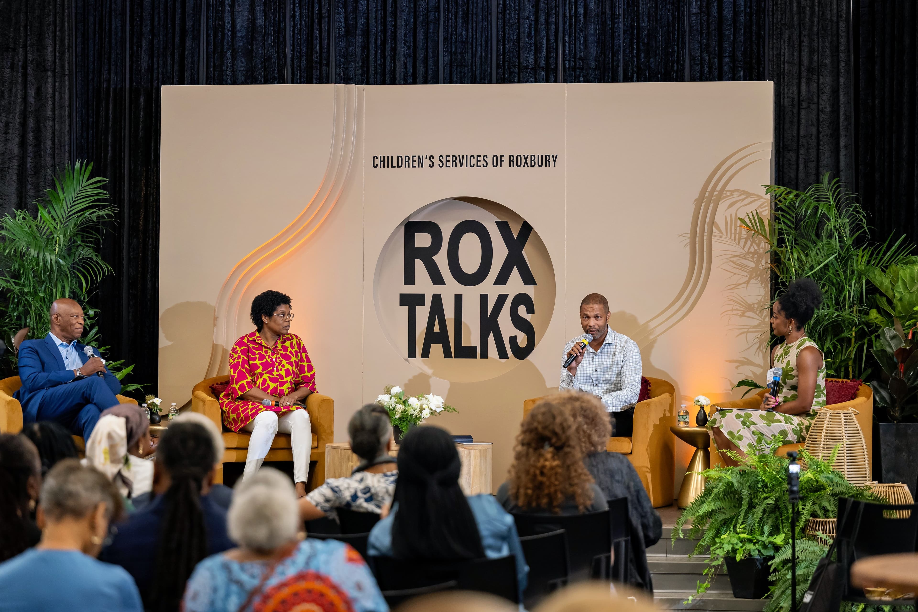Four people seated on stage in yellow armchairs, one speaking into a microphone, a large circular backdrop behind them.