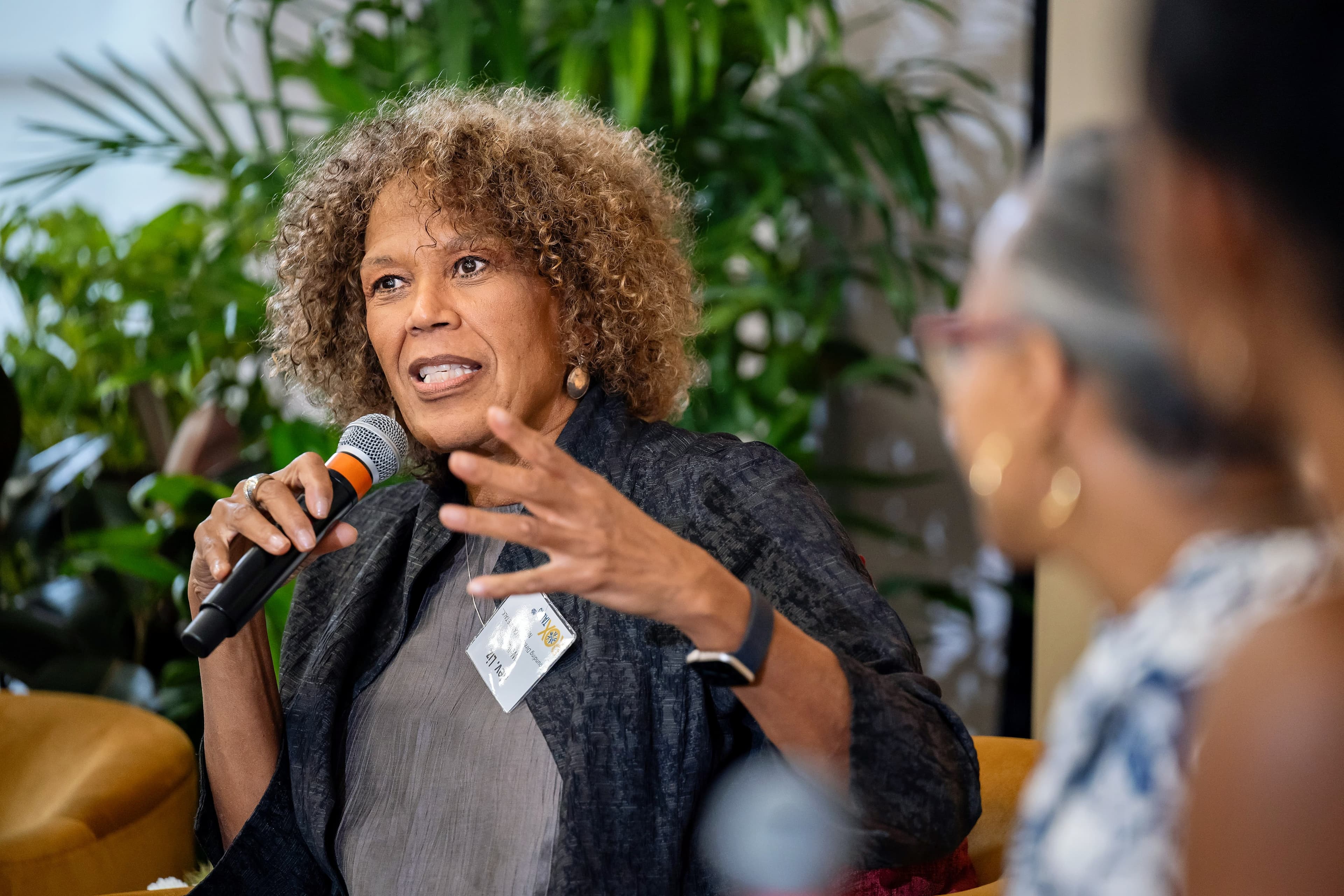 Woman seated and gesturing with a microphone in hand, another woman blurred in the background, tropical plants behind her.