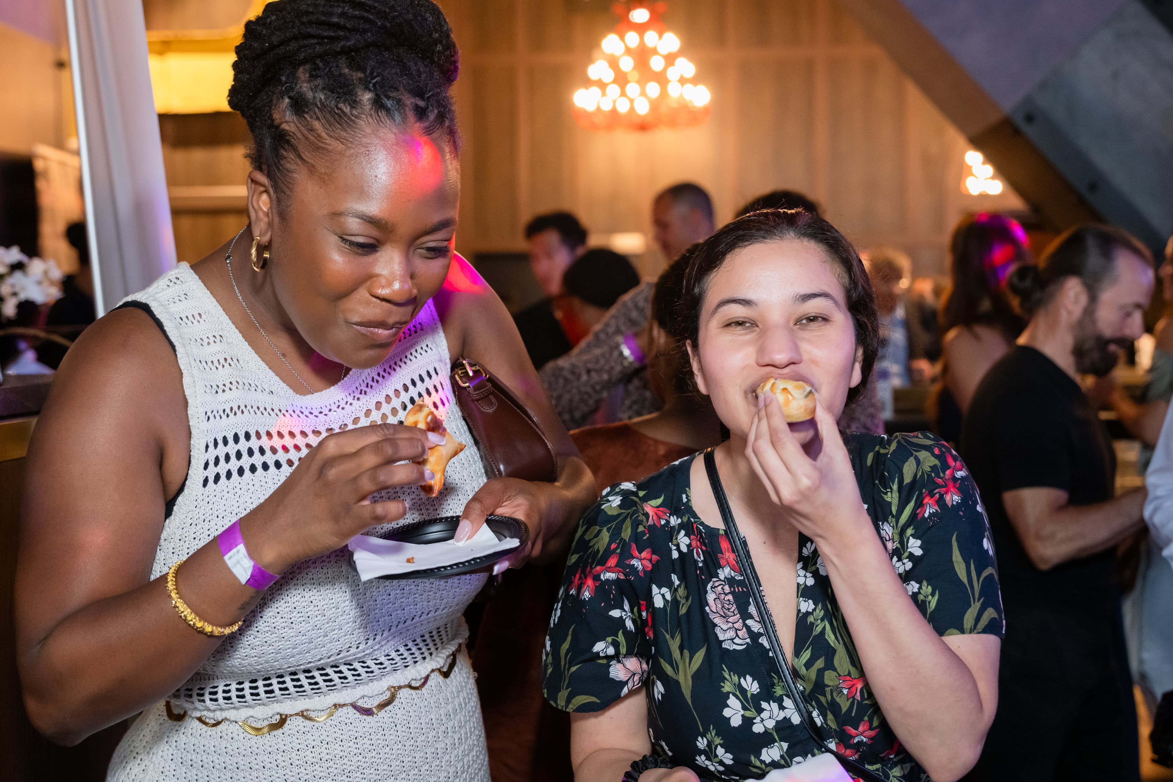 Two women eating appetizers together, one in a white crochet top holding a small plate, warm pendant lighting behind them.