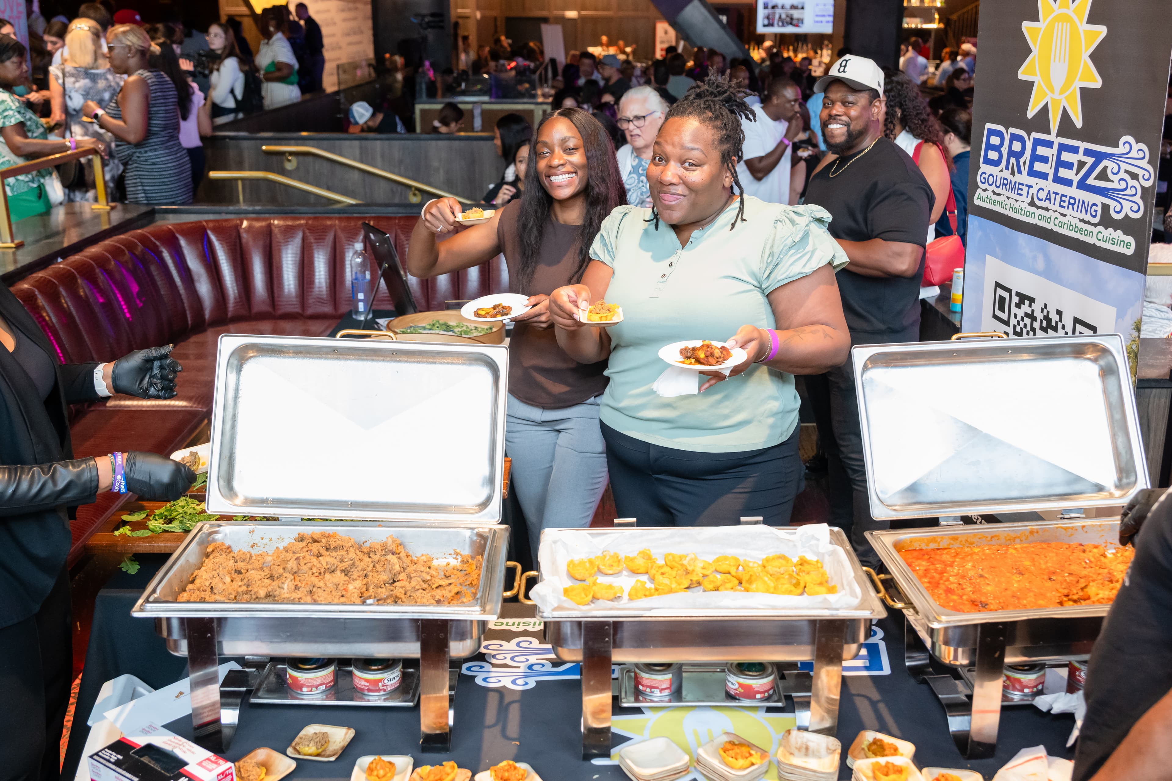 Two women and a man holding small plates of food in front of catering trays, a gourmet catering sign visible beside them.