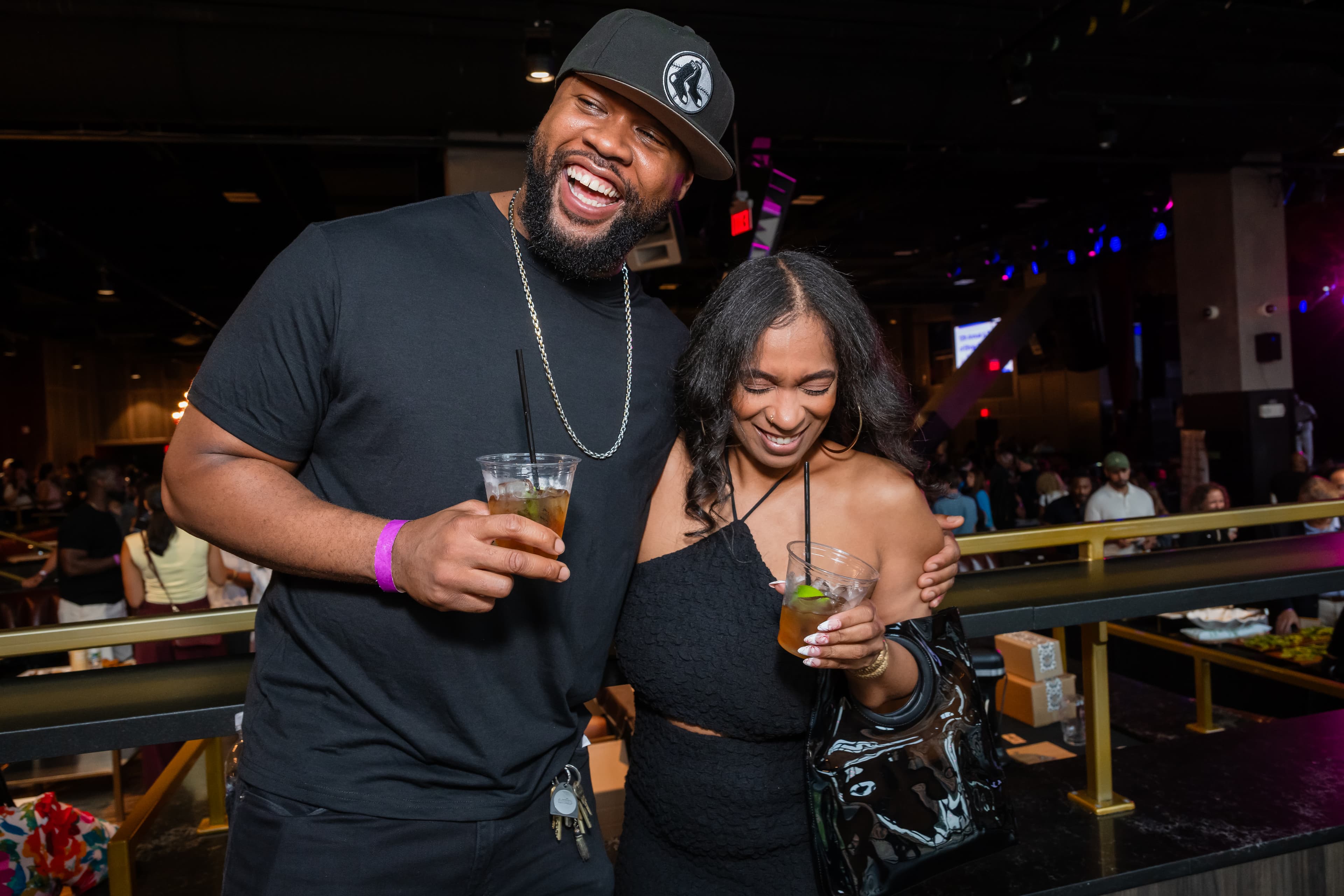 Man in a black cap laughing with his arm around a woman in a black dress, both holding drinks, string lights behind them.
