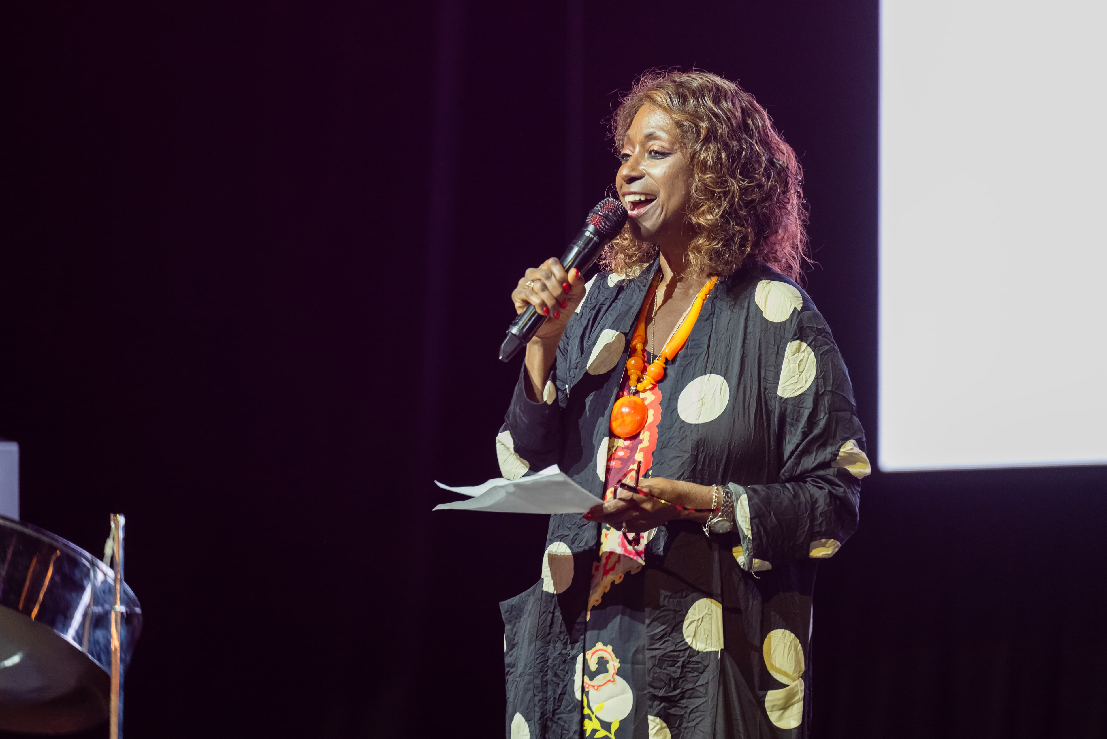 Woman in a black and cream polka-dot dress speaking into a microphone on stage, beaded necklace, dark curtain backdrop.